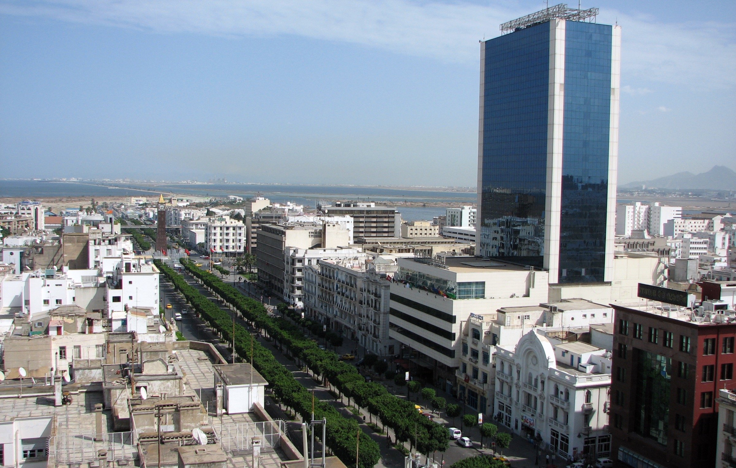L'image montre un paysage urbain, probablement d'une ville côtière avec une vue panoramique. On peut voir de grands bâtiments modernes, dont une tour en verre qui se dresse au centre. La rue est bordée d'arbres et d'immeubles, créant une ambiance sympathique. À l'arrière-plan, on aperçoit l'océan, suggérant que la ville est située près de la mer. Le ciel est dégagé avec quelques nuages, indiquant une belle journée.