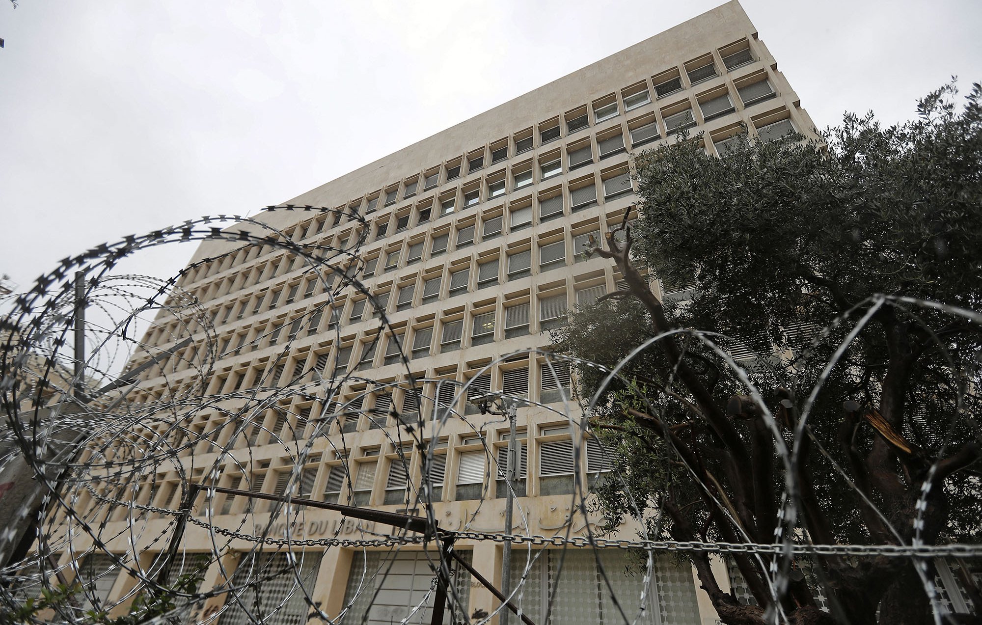 الصورة تظهر مبنى عالٍ يظهر بشكل معماري حديث، ويتواجد في المقدمة سياج من الأسلاك الشائكة. السماء فوق المبنى ملبدة بالغيوم، مما يوحي بجو قاتم. تحيط بالمبنى بعض الأشجار، مما يضيف لمسة خضراء في المشهد. تعكس الصورة شعورًا بالأمان أو القيود، نظراً للسياج الذي يحيط بالمكان.