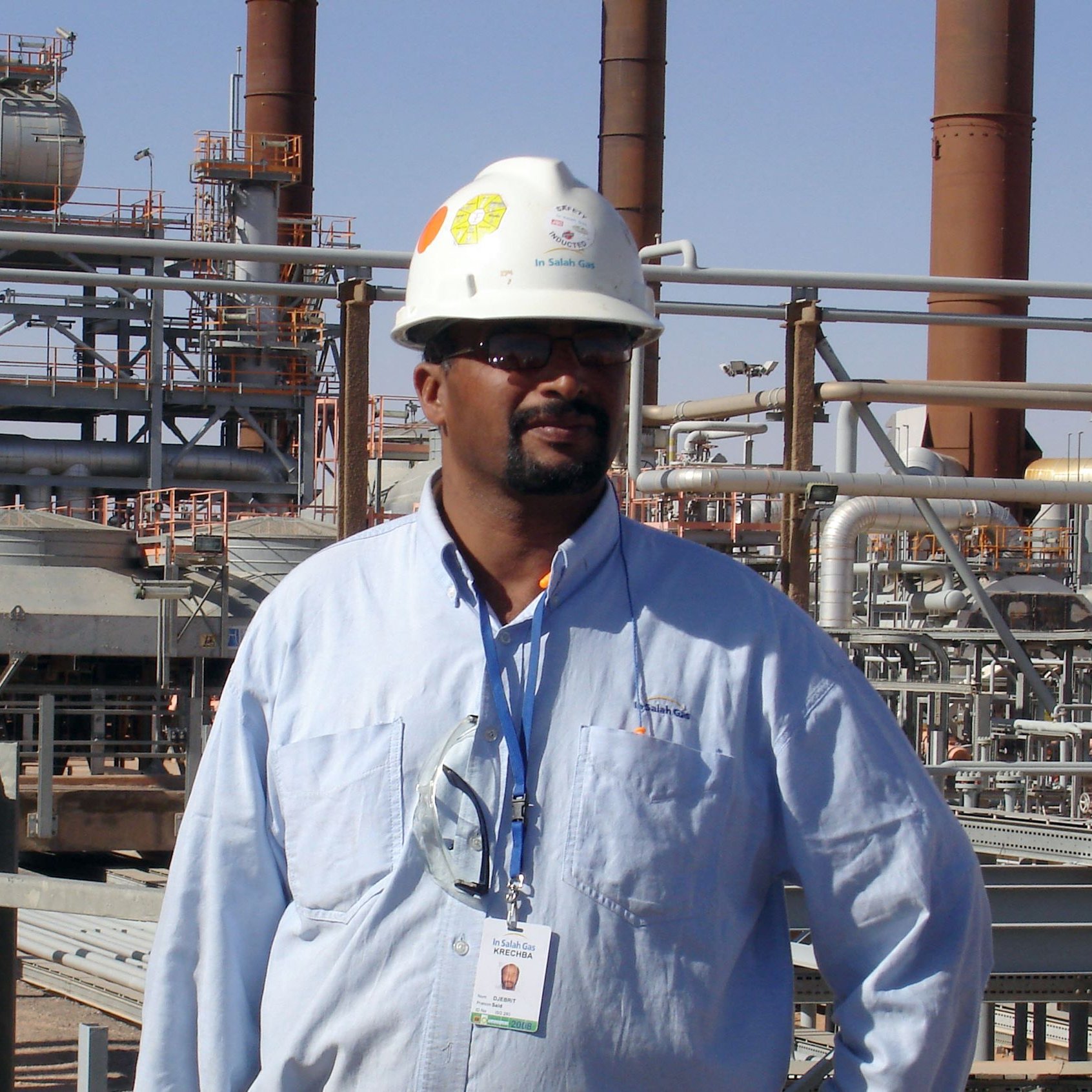 L'immagine mostra un uomo che indossa un casco di sicurezza bianco e una camicia azzurra. Si trova in un ambiente industriale, con impianti e tubazioni sullo sfondo. L'atmosfera sembra quella di un sito di produzione o di una raffineria, con diverse strutture metalliche e torri visibili. L'uomo appare fiducioso e professionale mentre si pone di fronte all'obiettivo.