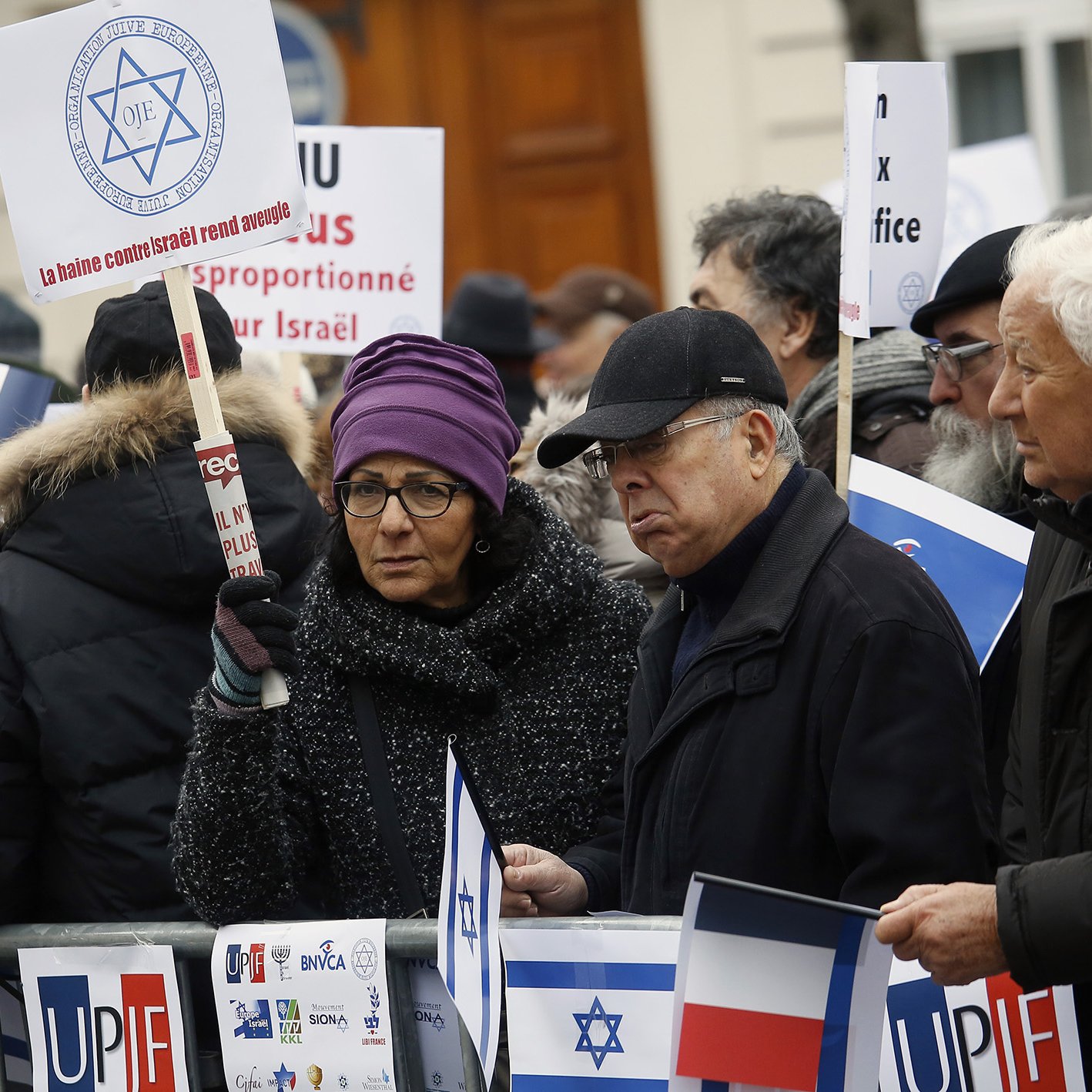 La imagen muestra a un grupo de personas participando en una manifestación. Los manifestantes sostienen pancartas y banderas, muchas de ellas con símbolos relacionados con Israel y Francia. Los asistentes varían en edad y están vestidos de manera variada, algunos con abrigos y gorros, lo que sugiere que es un día frío. El ambiente parece ser de protesta, con mensajes que expresan opiniones sobre el conflicto en Israel.