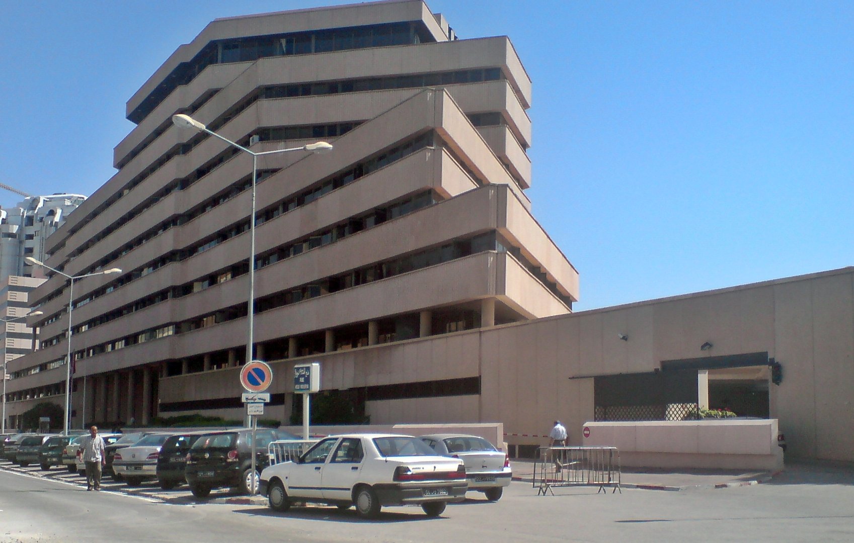 L'image montre un bâtiment moderne à plusieurs étages avec une architecture angulaire et des balcons en saillie. La façade est en béton, avec des couleurs neutres. On peut voir des voitures garées le long de la route, et il semble y avoir des piétons dans la zone. Le ciel est dégagé, ce qui donne une atmosphère ensoleillée à la scène. Le bâtiment dégage une impression de fonctionnalité et de modernité.