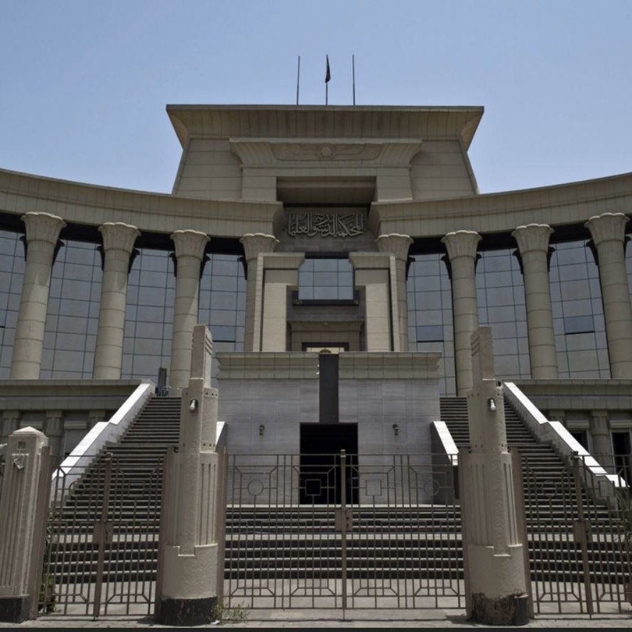 الصورة تظهر مبنى حكومي كبير يتميز بتصميم معماري حديث. يبرز في الصورة مدخل المبنى الذي يحتوي على مجموعة من السلالم والأعمدة الكبيرة. الواجهة تمتاز بالنوافذ الزجاجية العريضة التي تعكس الضوء، وتوجد أعلام مرفوعة فوق السطح. المنطقة المحيطة بالمبنى تحتوي على أشجار ونباتات تعطي جواً من الخضرة. يبدو الطقس مشمساً وواضحاً، مما يضفي على المشهد حيوية.