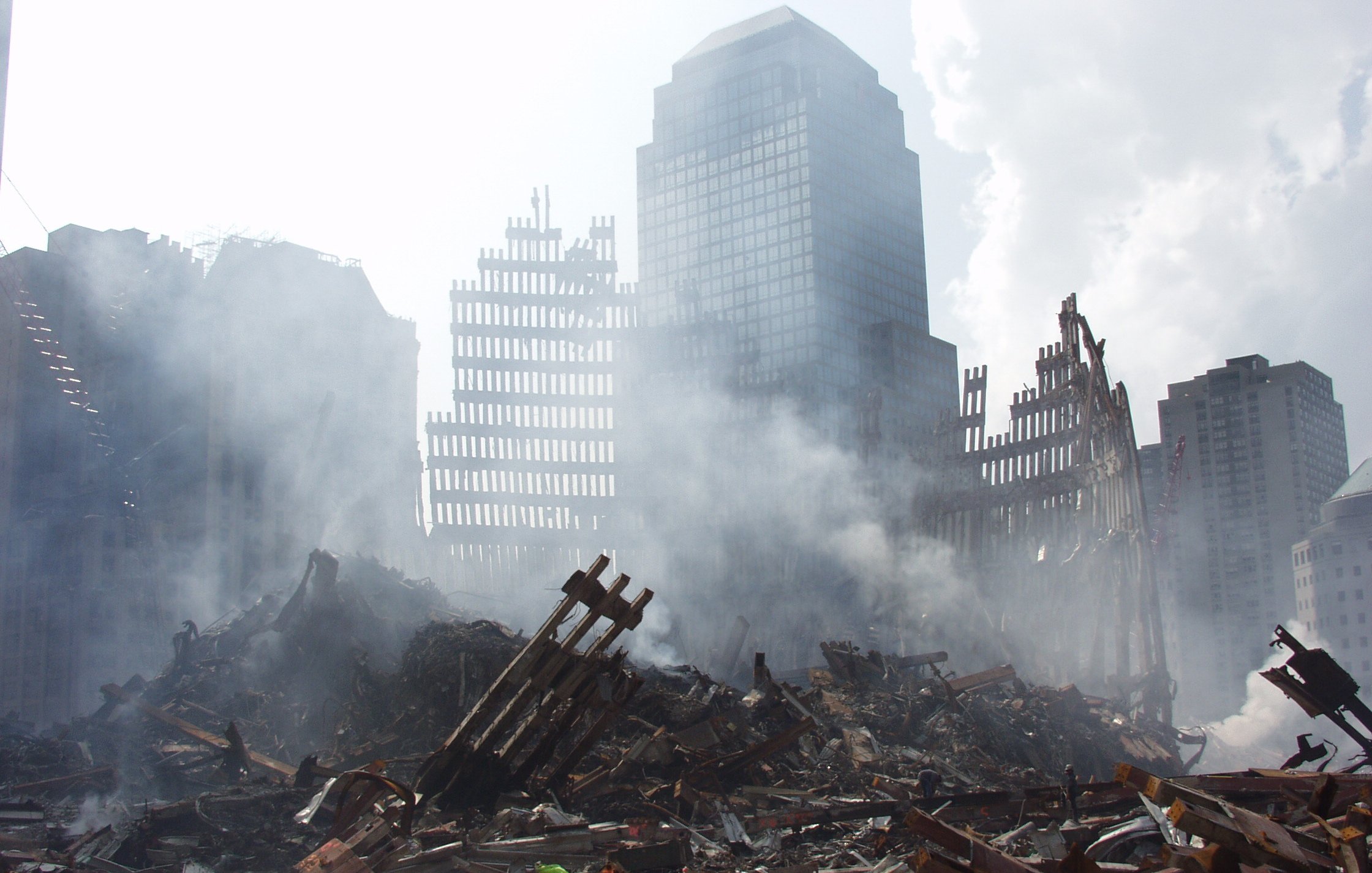 تصویر مربوط به یک صحنه خرابی و ویرانی است. در پس‌زمینه، ساختمان‌های بلند و ناتمام دیده می‌شوند که برخی از آنها به شدت آسیب‌دیده‌اند. در جلوی تصویر، خرابه‌هایی از سازه‌ها و تیرک‌های فلزی با دودی که از میان آنها بلند می‌شود، مشاهده می‌شود. حالت کلی تصویر نشان‌دهنده یک حادثه ویرانگر است و فضای ملتهب و غم‌انگیزی را ایجاد می‌کند.