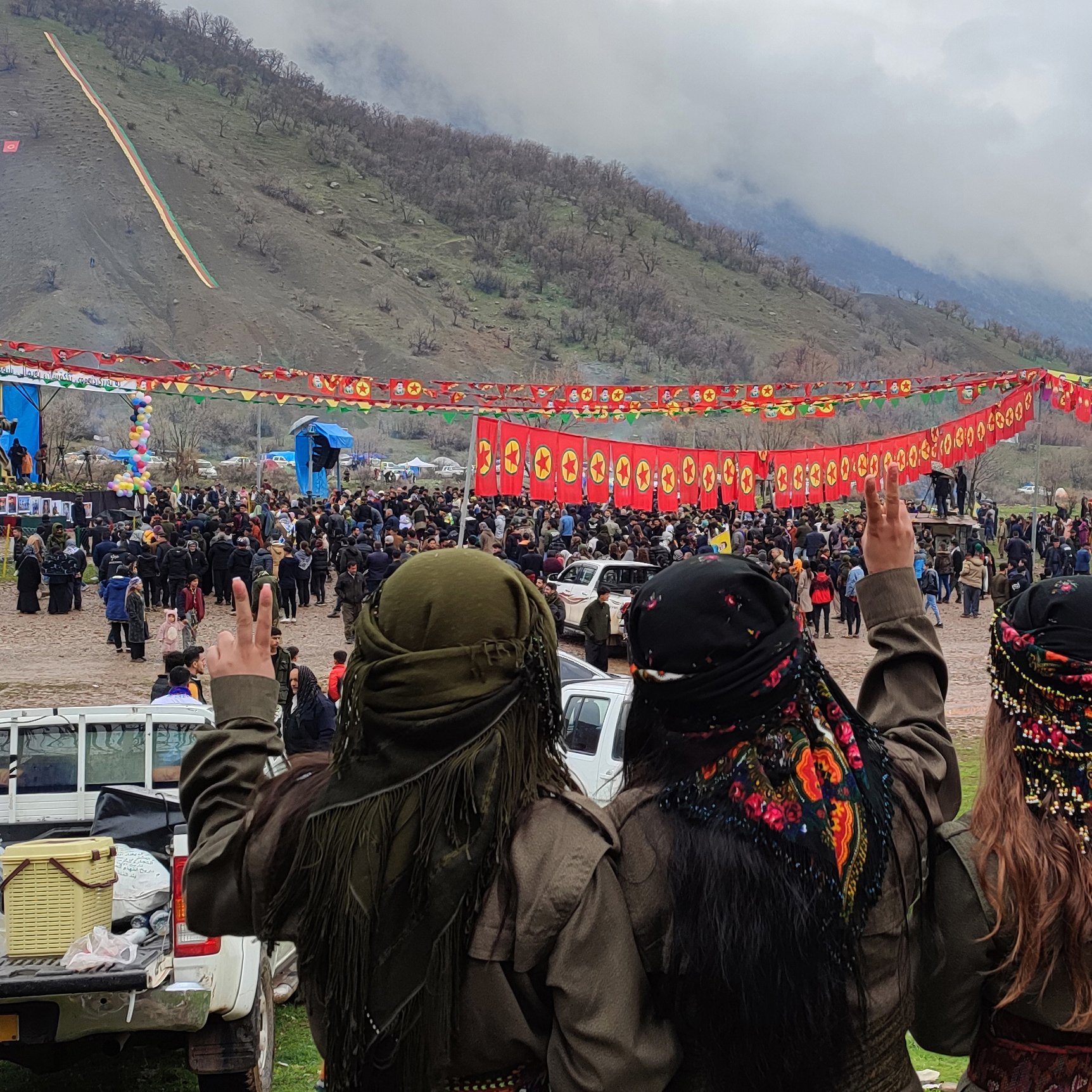 در این تصویر، چند نفر از پشت به دوربین ایستاده‌اند و در حال ارسال علامت پیروزی با دستان خود هستند. آنها لباس‌های محلی و رنگارنگی به تن دارند و با پوشش‌های سنتی روی سرشان تزئین شده‌اند. در پس‌زمینه، جمعیتی بزرگ در حال جشن و رقص در یک فضای باز دیده می‌شود. فضای اطراف دارای تزیینات رنگارنگ و پرچم‌هایی با طراحی‌های سنتی است. کوه‌ها و آسمان ابری در پس‌زمینه قرار دارند که حس طبیعت و جشنواره را تقویت می‌کند.