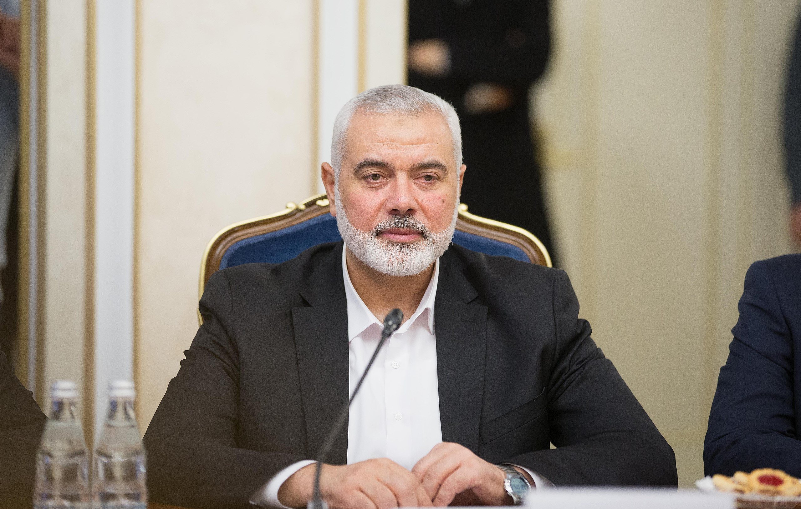 در تصویر، مردی با ریش خاکستری و موهای کوتاه دیده می‌شود که در حال نشستن در یک جلسه یا کنفرانس است. او ظاهری جدی دارد و یک کت تیره به تن دارد. بر روی میز مقابل او بطری‌های آب و تعدادی خوراکی دیده می‌شود. فضای اطراف به نظر رسمی و مرتبط با مذاکرات یا بحث‌های مهم می‌رسد.