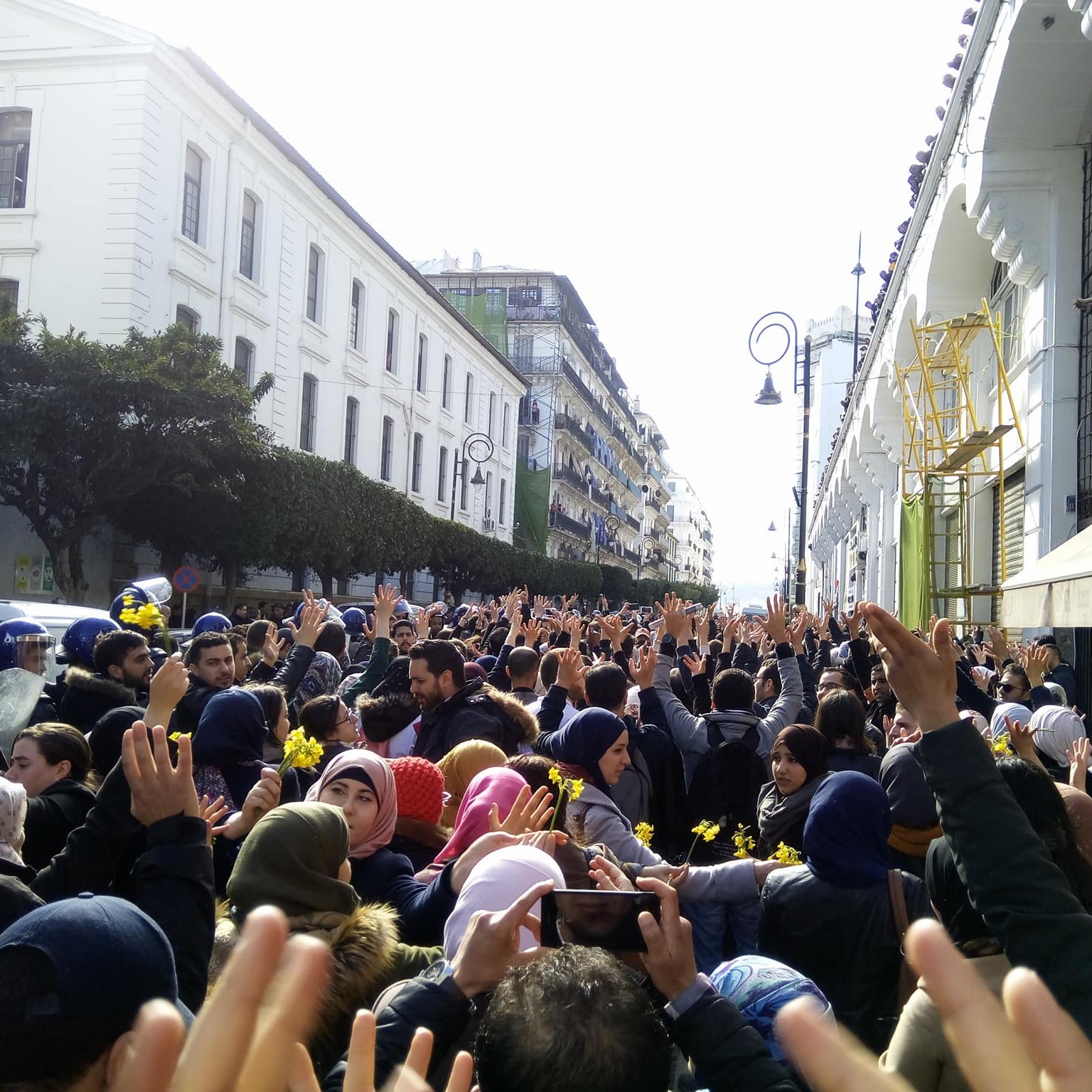 The image depicts a large crowd of people gathered in an urban setting, possibly during a protest or demonstration. Many individuals are raising their hands, and a sense of unity and collective action is present. The atmosphere appears to be lively, and the crowd is surrounded by buildings, suggesting a city environment. Some police presence is visible, indicating that the gathering may be significant or organized and possibly involves a call for change or social issues. The crowd includes diverse individuals, wearing a range of clothing styles, with some wearing headscarves.