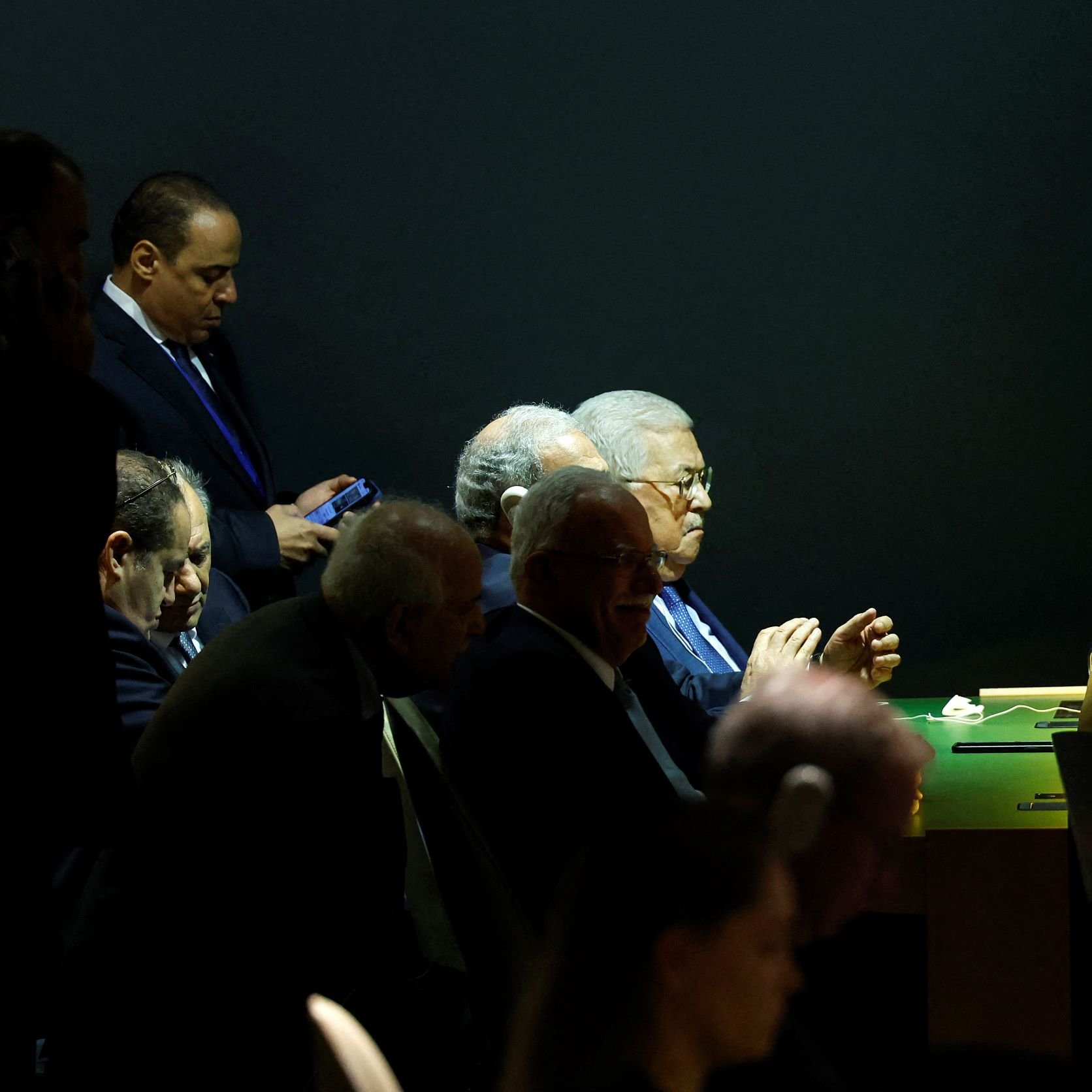 در این تصویر، گروهی از مردان در یک محیط تاریک نشسته‌اند. برخی از آنها در حال صحبت هستند و تمرکز دارند. در میان آنها، فردی با عینک و موهای خاکستری توجه بیشتری را جلب می‌کند. یکی از افراد نیز در حال استفاده از گوشی موبایل خود است. فضای اطراف شامل نورهای ملایمی است که بر روی صحنه یا میز قرار دارد. این تصویر فضایی رسمی و جدی را القا می‌کند.