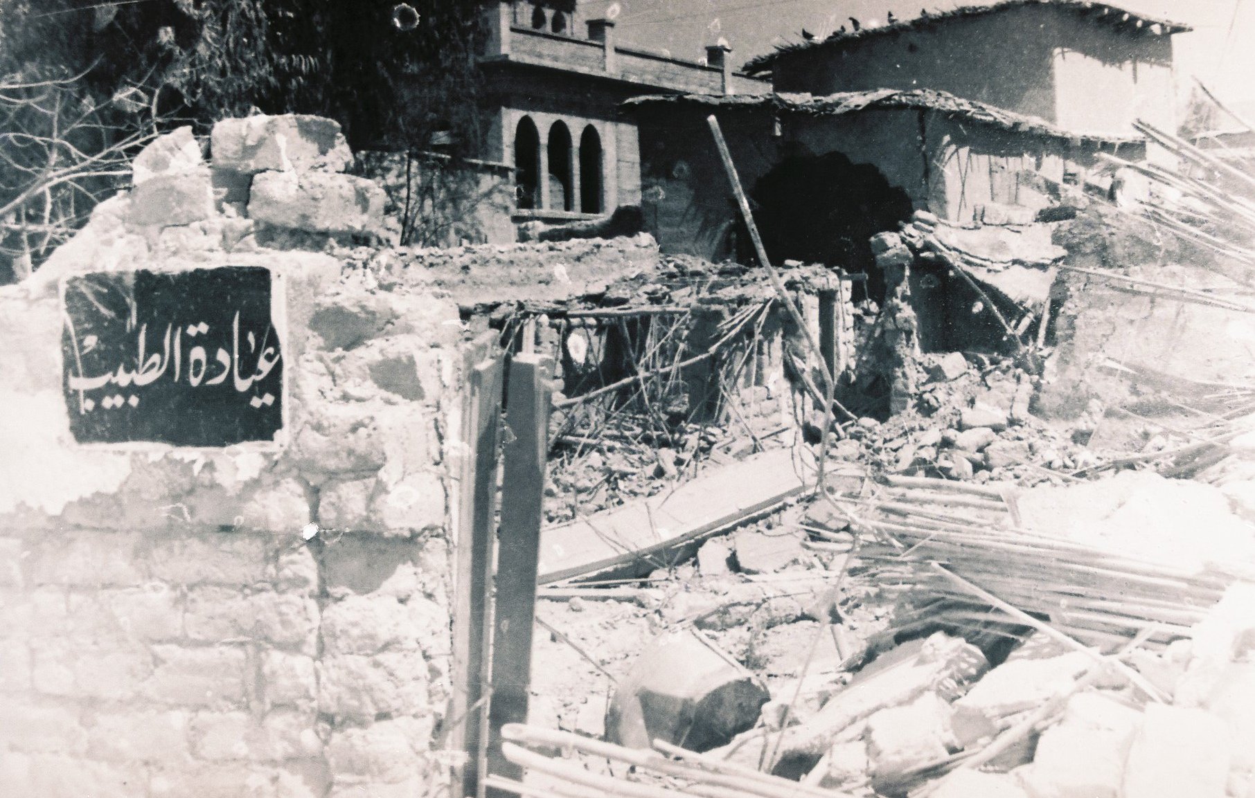 L'image montre une scène de destruction, avec des bâtiments en ruines. On peut apercevoir un mur effondré, laissant entrevoir des débris et des structures endommagées. À côté des débris, il y a une plaque signalétique en arabe qui semble indiquer un lieu ou un bâtiment. L'atmosphère générale de la photo suggère un environnement dévasté, probablement à la suite d'un conflit ou d'un désastre.