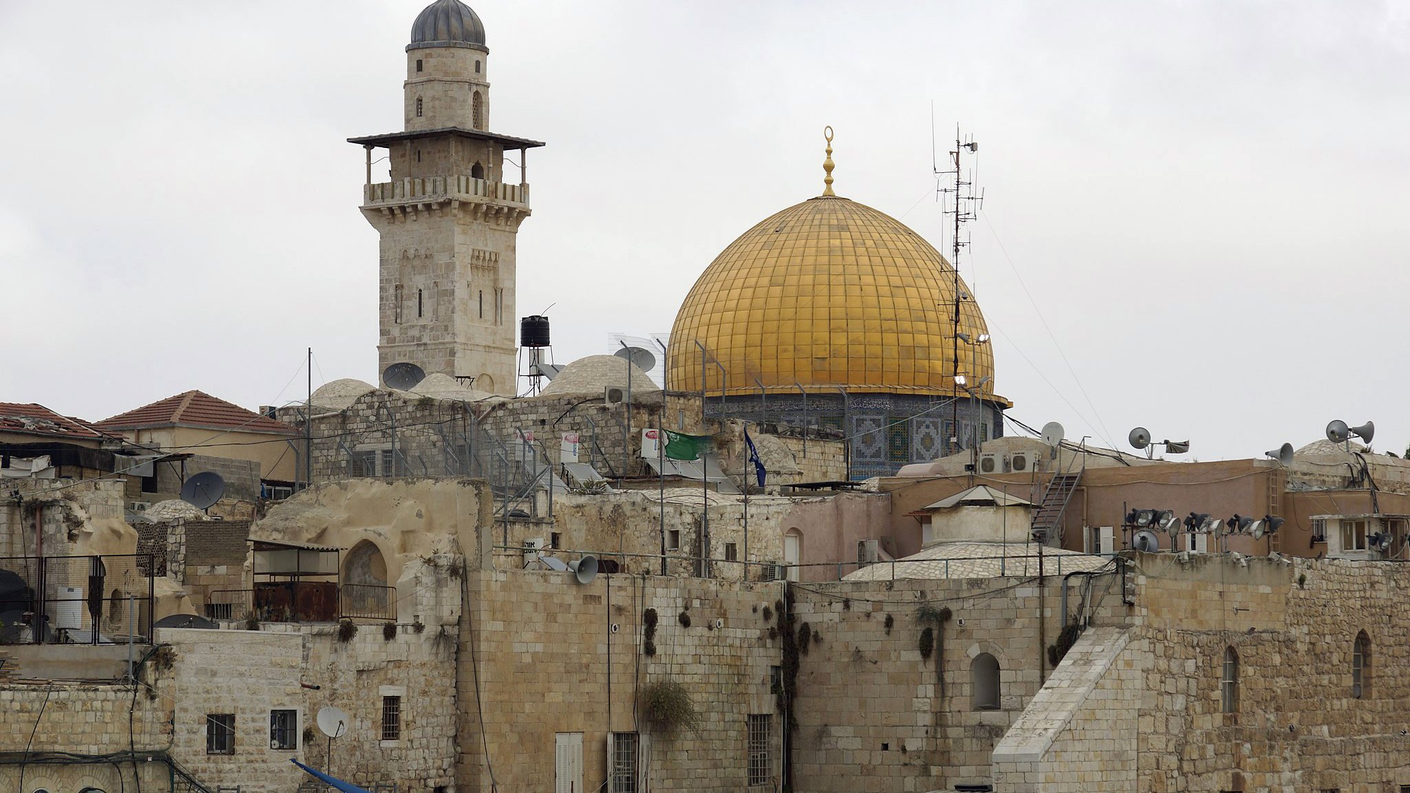 تظهر الصورة مشهداً لمدينة القدس، حيث يتوسطها المسجد الأقصى، الذي يبرز بقبة ذهبية مميزة. بجانب القبة، يمكن رؤية مئذنة ترتفع عالياً، وهناك بعض المباني التاريخية ذات الحجر القديم. كما يظهر في الصورة وجود أجهزة استقبال الأقمار الصناعية على أسطح المباني، مما يعكس مزيجاً من التاريخ والحداثة في هذه المنطقة. السماء تبدو غائمة، مما يضفي على المشهد جوّاً من السكون.