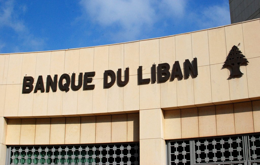 تظهر الصورة واجهة مؤسسة مالية تحمل اسم "بنك لبنان" بالخط العريض. الكتابة باللون الأسود على خلفية فاتحة، وتوجد شجرة الأرز، وهي رمز وطني للبنان، بجانب الاسم. السماء زرقاء مع بعض السحب، مما يعطي انطباعًا بوجود يوم مشمس. يبدو أن الجزئية المعروضة من المبنى حديثة ومرتبة.