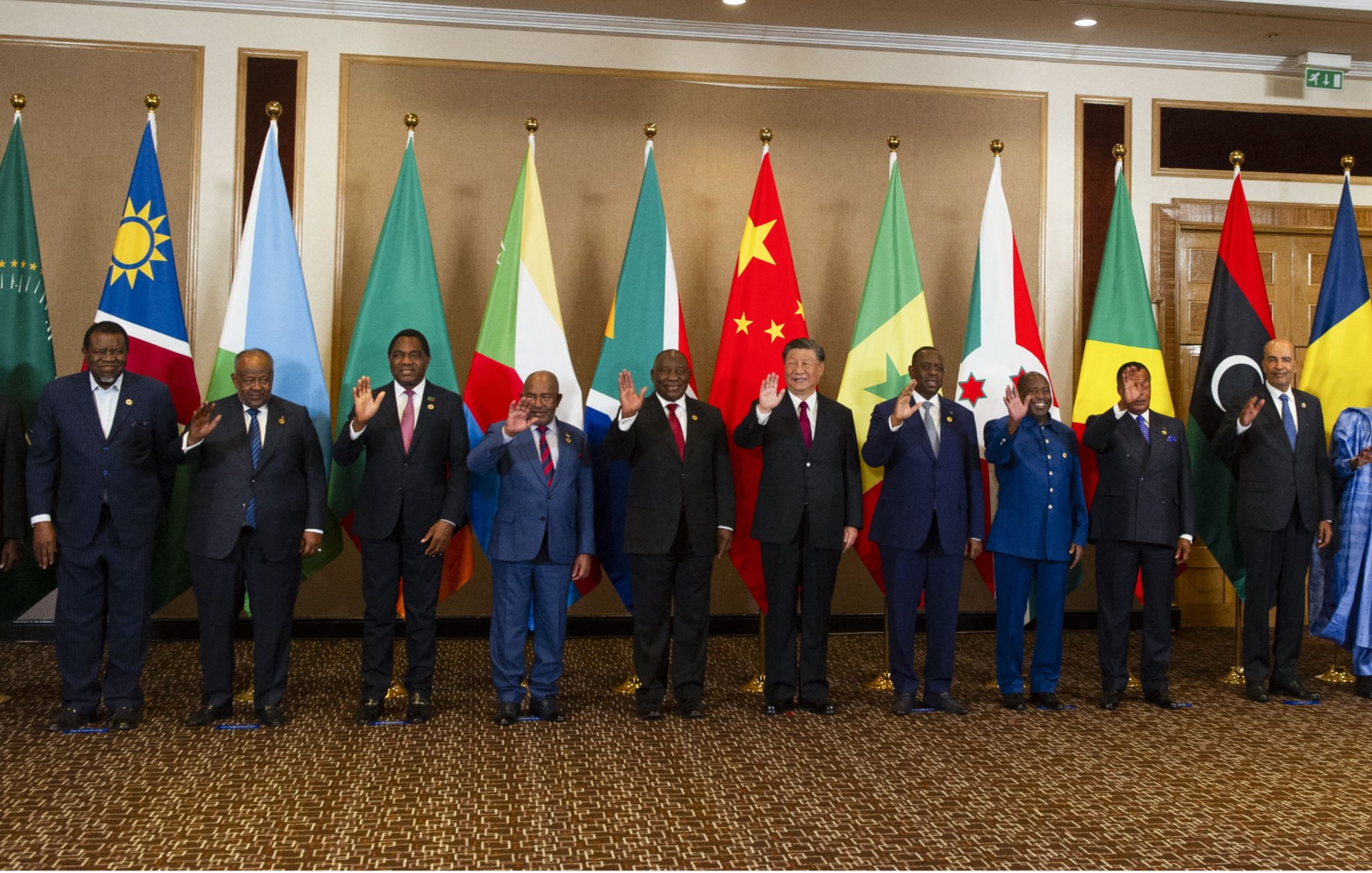 L'image montre un groupe de personnes debout en rangée, saluant. Elles se trouvent devant une série de drapeaux représentant différents pays. Les personnes portent des costumes formels et semblent participer à un événement diplomatique ou à un sommet international. Les drapeaux en arrière-plan incluent ceux de l'Union africaine, de la Chine, ainsi que d'autres nations. L'ambiance générale semble conviviale et respectueuse.