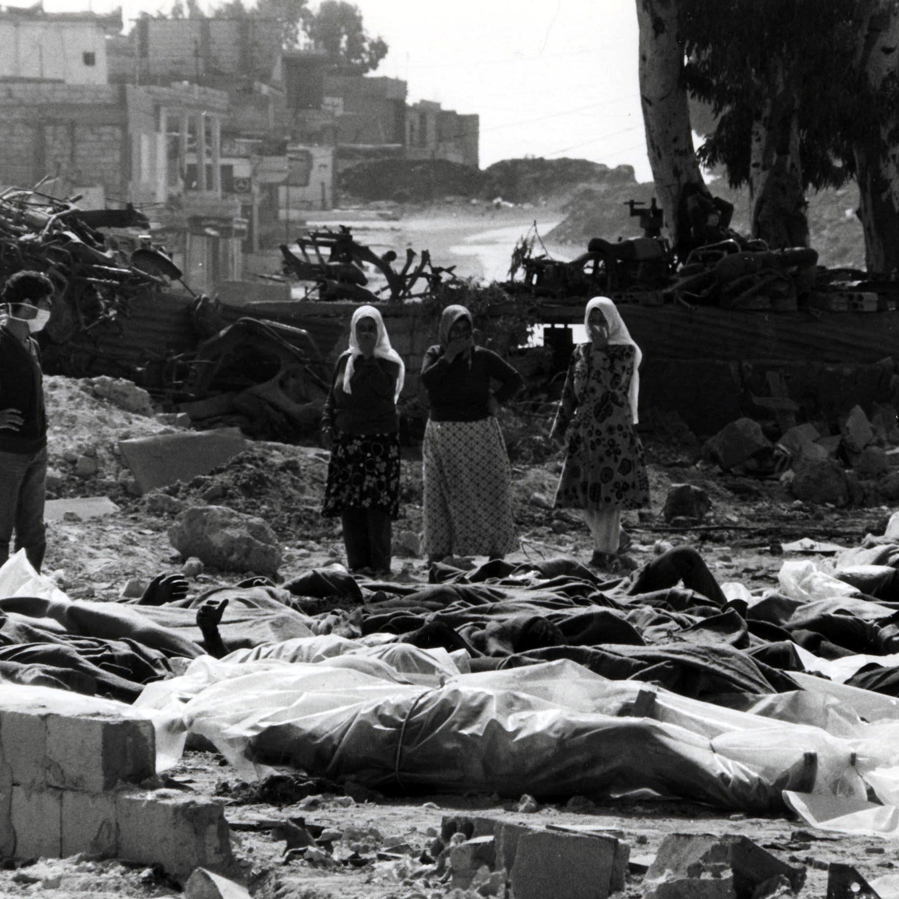 این تصویر نمایی غم‌انگیز و دل‌خراش از یک صحنهٔ بعد از بحران یا جنگ است. در جلوی تصویر، چندین جسد که با پارچه‌ای پوشیده شده‌اند، قرار دارند. در پس‌زمینه، گروهی از افراد حاضرند که به این صحنه نگاه می‌کنند، برخی از آنها اقدام به پوشاندن دهان و بینی خود کرده‌اند. درختان و ساختمان‌های ویرانه در پشت زمینه به چشم می‌خورند، که حاکی از تخریب و ویرانی است. جو کلی تصویر شعاری از درد و مصیبت را منتقل می‌کند.