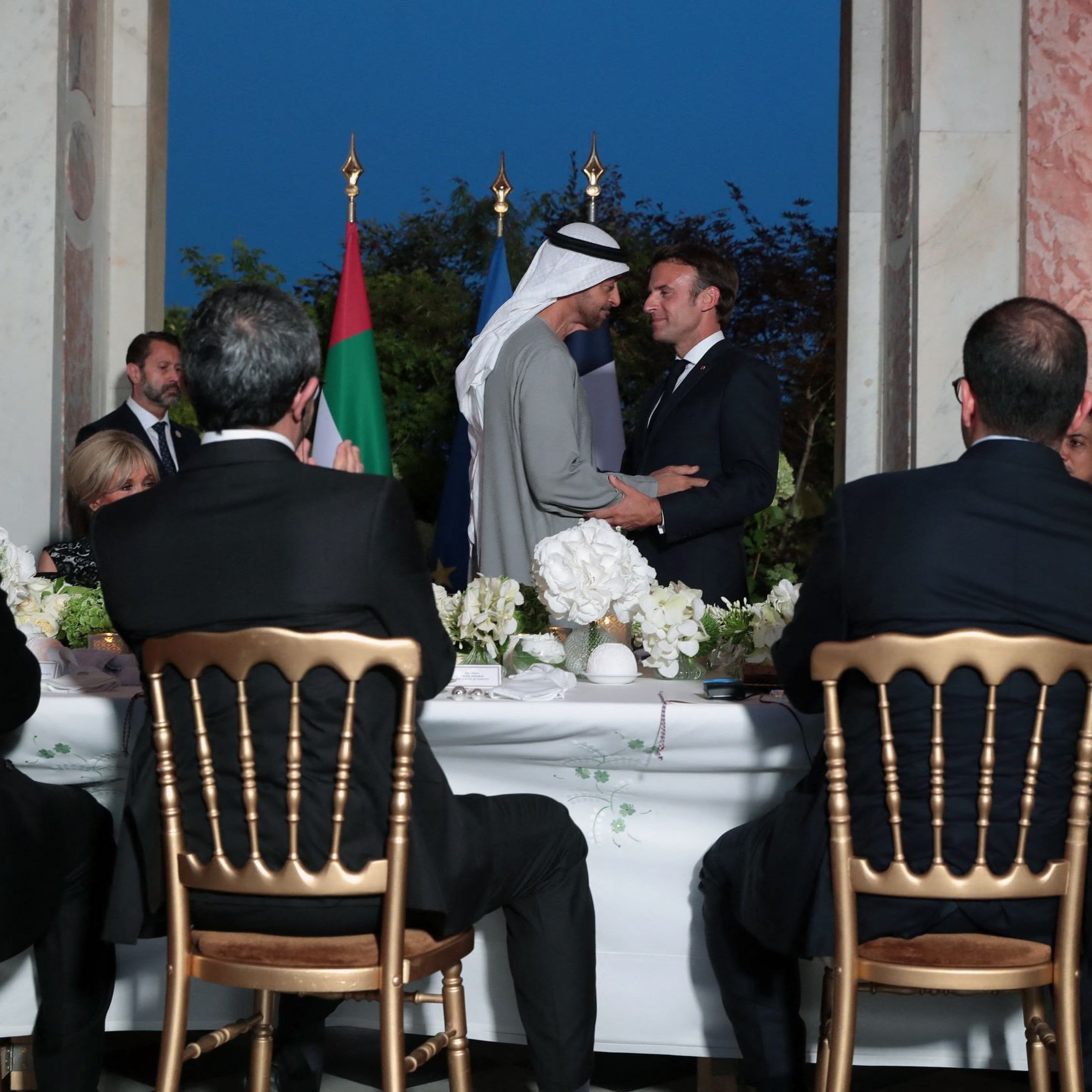 The image depicts a formal gathering, likely a diplomatic event or dinner. In the foreground, several individuals are seated at a long table, facing away from the camera. They appear to be engaged in conversation or listening attentively. In the background, two men are shaking hands or exchanging greetings, with flags visible nearby, suggesting a moment of diplomacy or cooperation. The setting features elegant decor, with floral arrangements on the table and a softly lit ambiance, indicating an evening event.