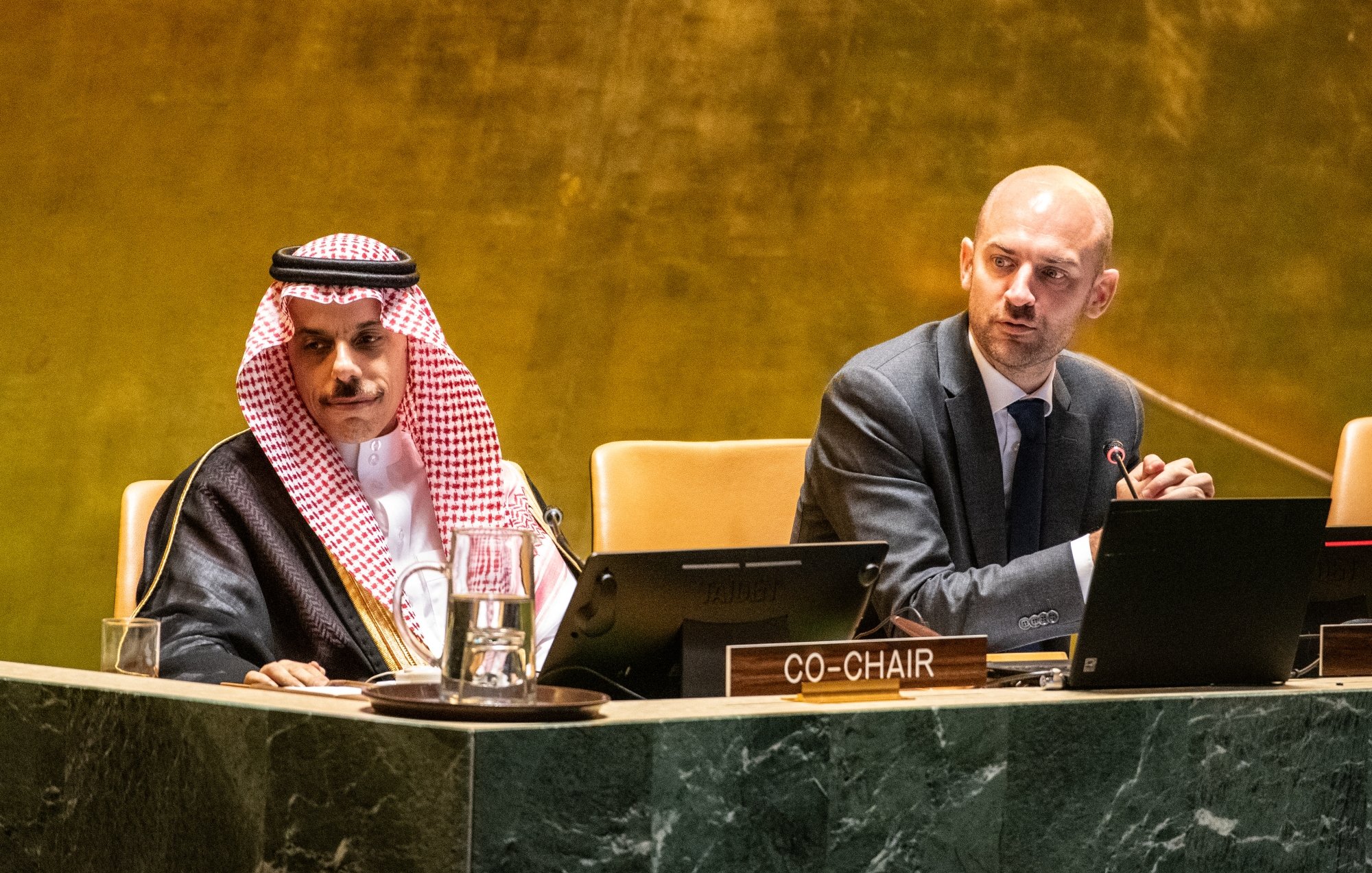 Two men at a conference table, one in traditional attire, discussing on a stage with microphones.