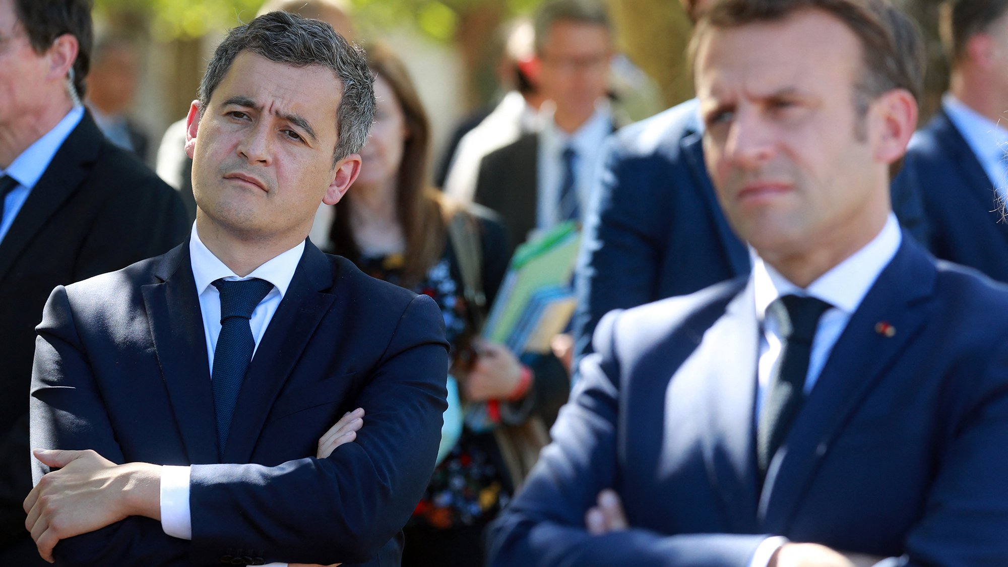 L'image montre deux hommes en costume, l'un avec les bras croisés et l'air pensif, et l'autre légèrement en avant, observant. Ils semblent participent à un événement public, entourés d'autres personnes en arrière-plan, dans un cadre extérieur. Les expressions faciales suggèrent qu'ils écoutent attentivement ou réfléchissent à ce qui se passe.