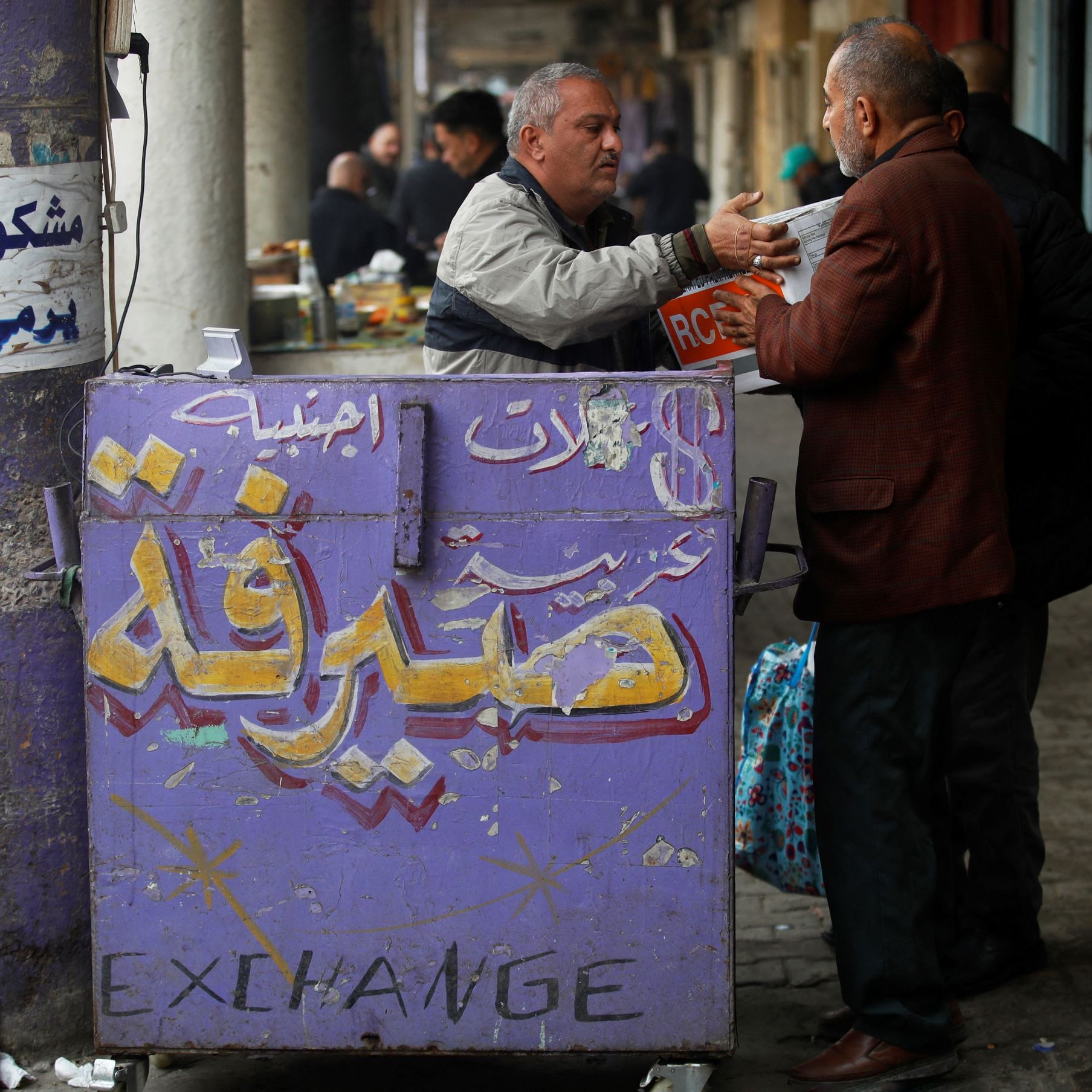 L'immagine mostra una scena di vita quotidiana in un mercato. Ci sono due uomini che interagiscono; uno sembra stia scambiando qualcosa con l'altro, mentre un'insegna colorata in arabo e inglese, che indica un'agenzia di cambio, è visibile in primo piano. Lo sfondo è caratterizzato da colonne e da un'atmosfera vivace tipica dei mercati affollati. Le persone e le attività circostanti suggeriscono un ambiente commerciale attivo.