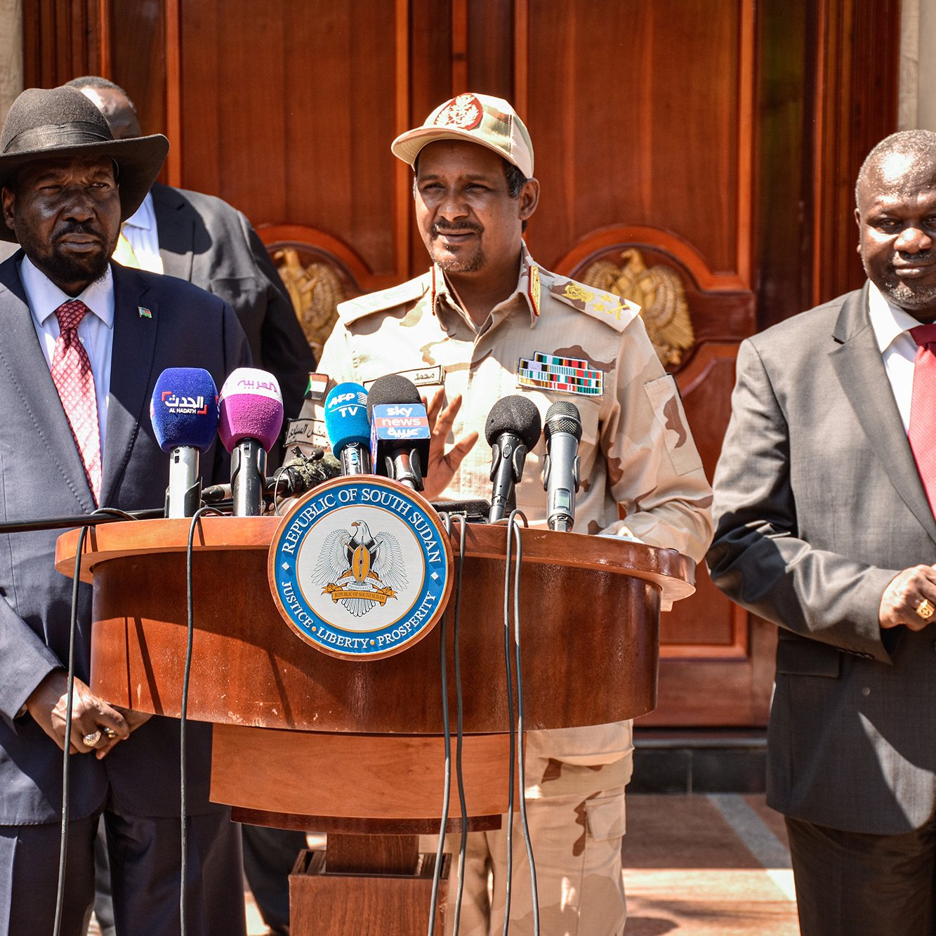 La imagen muestra a tres hombres de pie frente a un podio. El hombre a la izquierda lleva un sombrero y un traje negro, mientras que el del centro viste un uniforme militar con medallas y un gorra. El hombre a la derecha está vestido con un traje oscuro y parece estar atento a la conversación. Detrás de ellos, se puede ver un edificio con decoraciones arquitectónicas elaboradas. El ambiente parece formal, posiblemente en un contexto político o de conferencia de prensa.