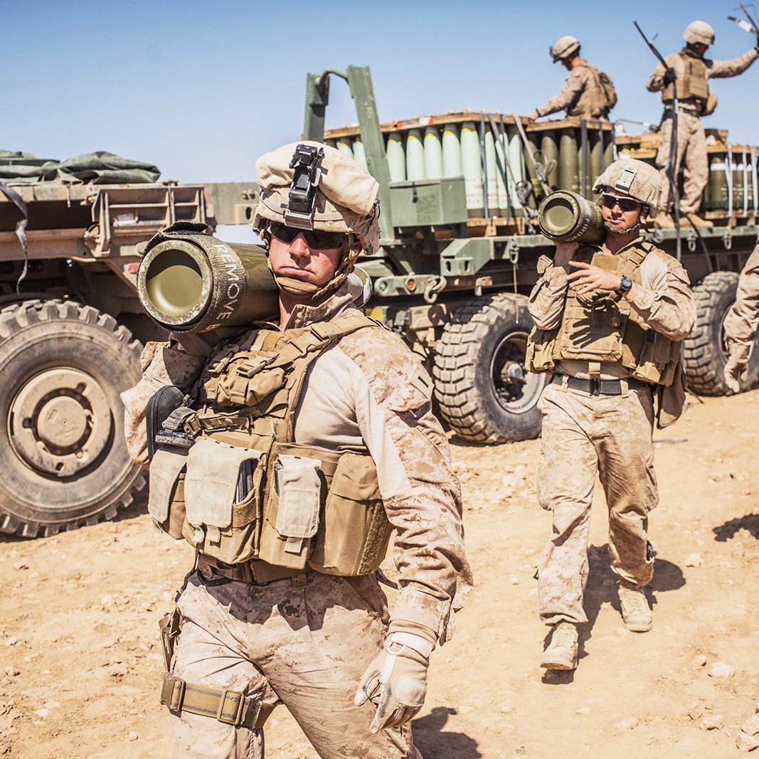 L'image montre des soldats en tenue militaire, probablement en opération. Certains d'entre eux portent des gilets tactiques et des casques, tandis qu'un soldat en avant tient un obus de mortier. En arrière-plan, on aperçoit un véhicule militaire avec une remorque et d'autres soldats s'affairant. Le cadre semble désertique, avec un sol sablonneux et un ciel dégagé. On ressent un climat de préparation et d'activité militaire.