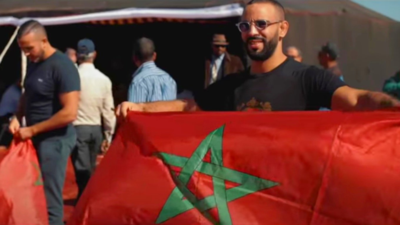 En la imagen se puede ver a un grupo de personas en un ambiente al aire libre, posiblemente en un evento o celebración. Un hombre sostiene una bandera de Marruecos, que es de color rojo con una estrella verde en el centro. Otros hombres también están presentes en el fondo, algunos sosteniendo banderas o participando en la actividad. La escena refleja un ambiente de camaradería y orgullo nacional.