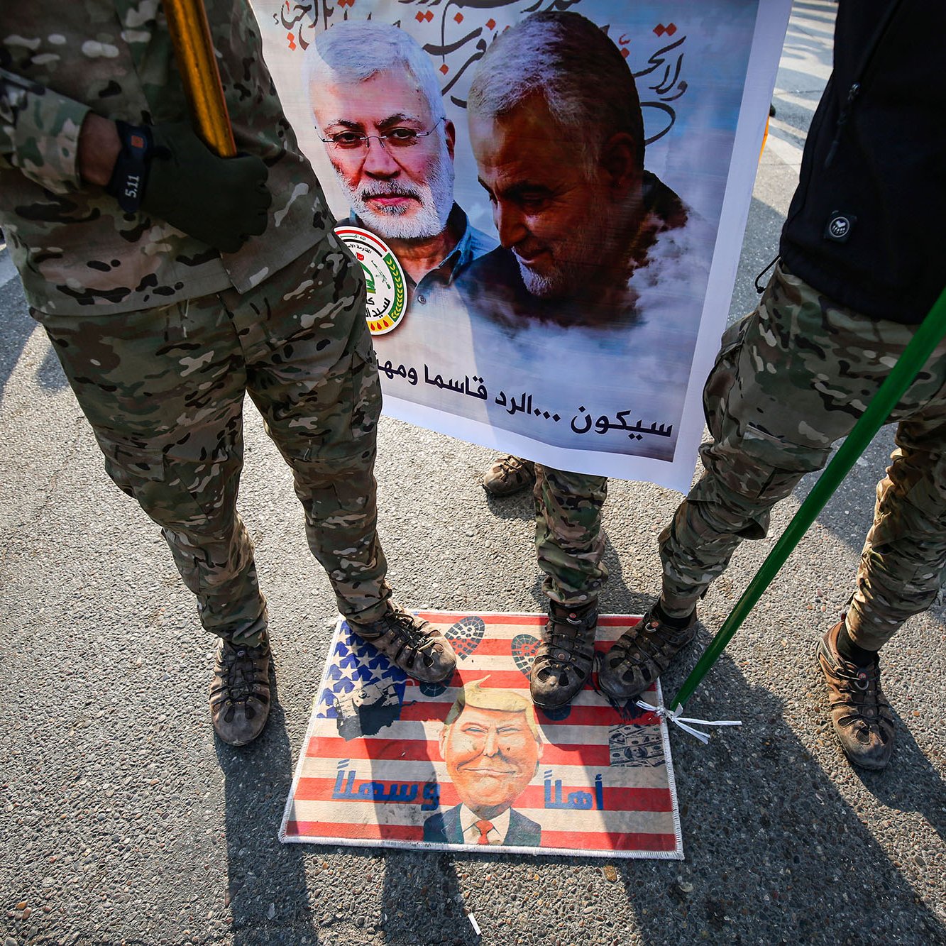 در این تصویر، تعدادی از افراد با لباس‌های نظامی و camo (رنگ‌های استتاری) در حال ایستادن هستند. آنها در دست خود عصا یا چوب‌هایی دارند. زیر پایشان تصویر یک پرچم آمریکاست که بر روی آن چهره‌ای در پایین مشاهده می‌شود. در پس‌زمینه، یک بنر بزرگ وجود دارد که شامل تصاویری از دو نفر است و متنی در مورد آنها نوشته شده است. فضایی حاکی از تظاهرات یا یک رویداد اجتماعی در حال شکل‌گیری است.