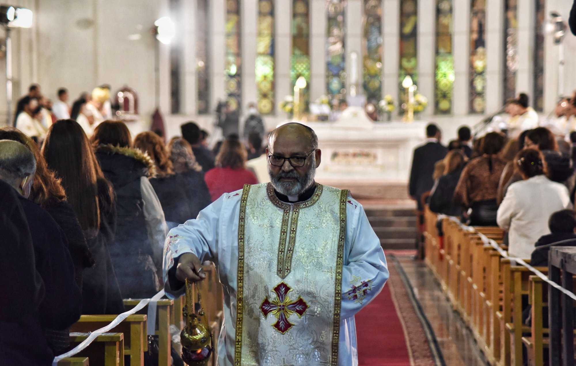 A priest in ornate vestments walks down an aisle in a church, surrounded by worshippers.