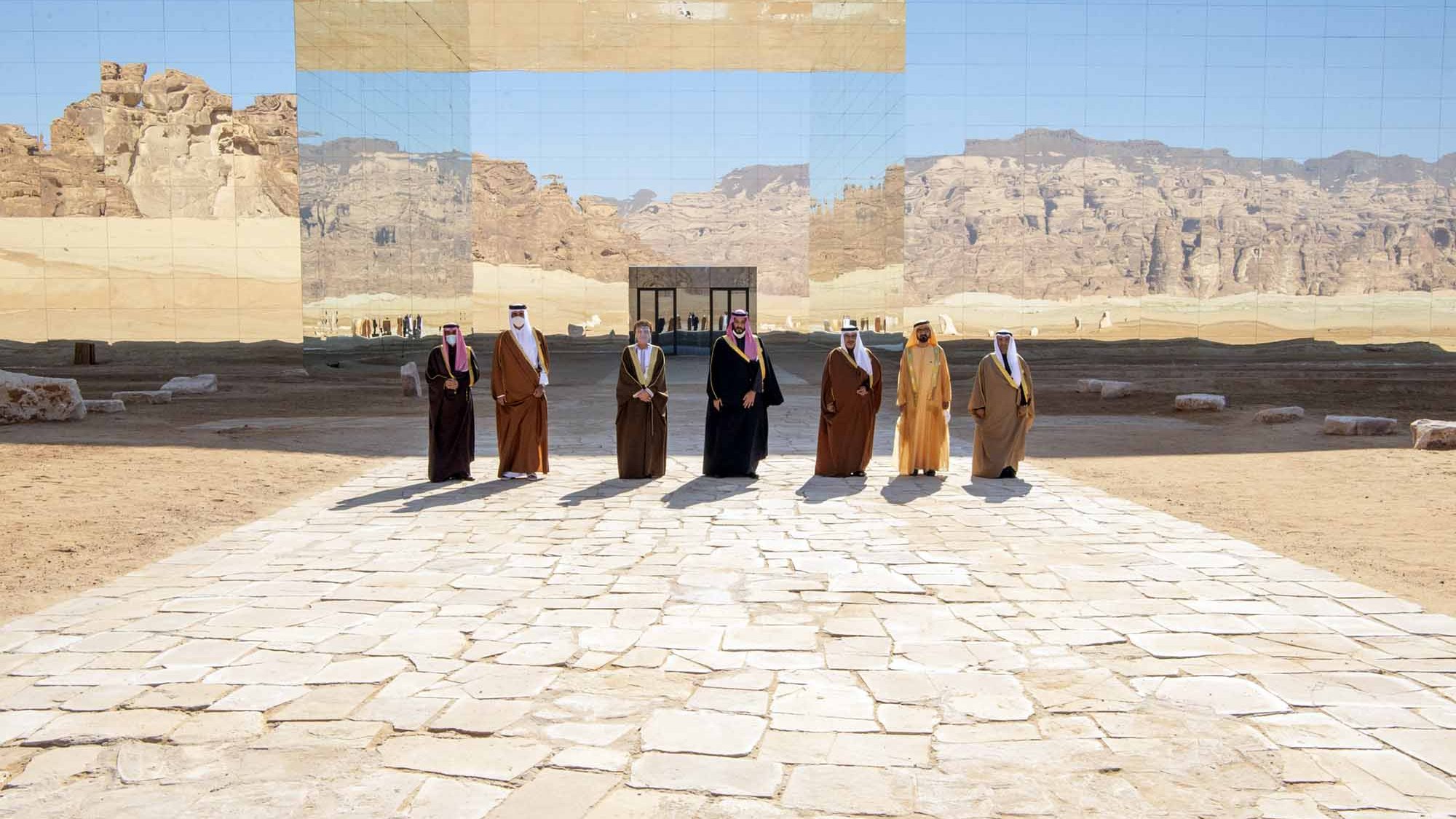 تظهر في الصورة مجموعة من الأشخاص يرتدون الزي التقليدي، ويقفوا على طريق مرصوف بالحجارة في منطقة صحراوية. خلفهم توجد جدران عاكسة تعكس المنظر الجبلي المحيط، مما يعطي شعوراً بالتواصل بين البشر والطبيعة. السماء زرقاء وصافية، مما يضيف جمالاً على المشهد.