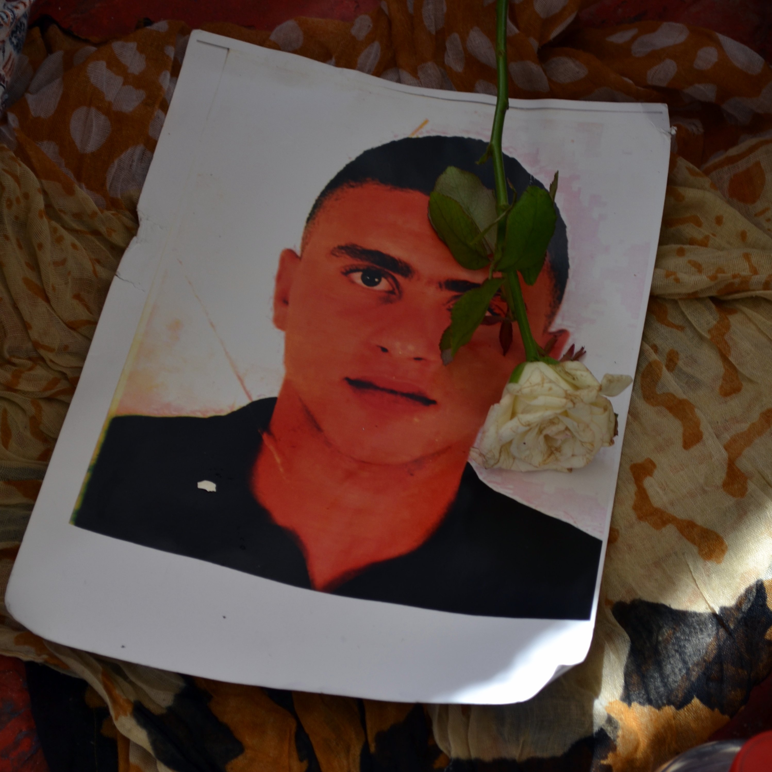 تظهر في الصورة ورقة مطبوعة تحتوي على صورة شاب، حيث يوضع عليها وردة بيضاء. الخلفية تبدو ملونة ومعقدة، مما يضيف جوًا من الحزن والحنين. الورقة قد تشير إلى ذكرى لهذا الشخص، والزهرة تعزز من مشاعر التأمل والاحترام.