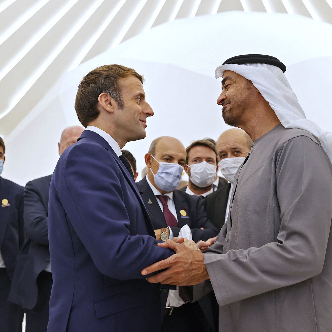 En la imagen se pueden ver a varias personas en un ambiente formal. Al frente, dos hombres están estrechando manos, uno de ellos viste un traje y el otro lleva una vestimenta tradicional árabe. Alrededor de ellos, hay más personas que observan la interacción, algunas de las cuales llevan mascarillas. El fondo muestra una arquitectura moderna y luminosa. La atmósfera es de cordialidad y diplomacia.