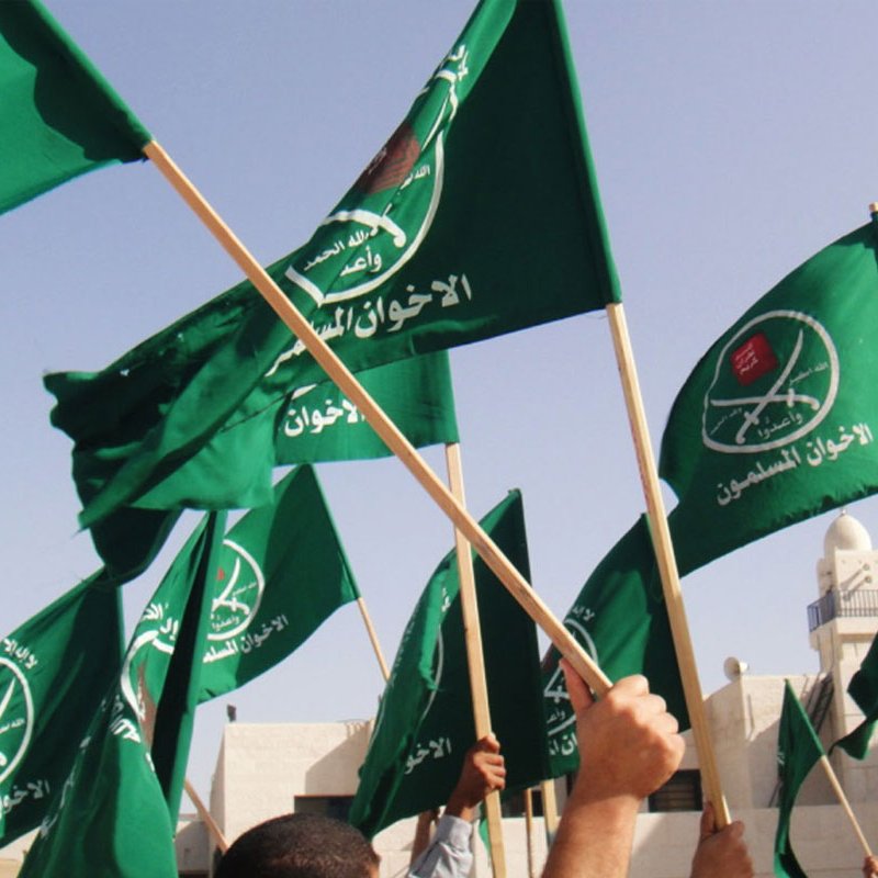 The image depicts a crowd of people holding up green flags. The flags feature a logo that includes Arabic text and a symbol, likely associated with a political or religious group. The scene appears to be one of a gathering or demonstration, with an emphasis on the flags being waved prominently. The background suggests an outdoor setting, possibly near a mosque or a public place.