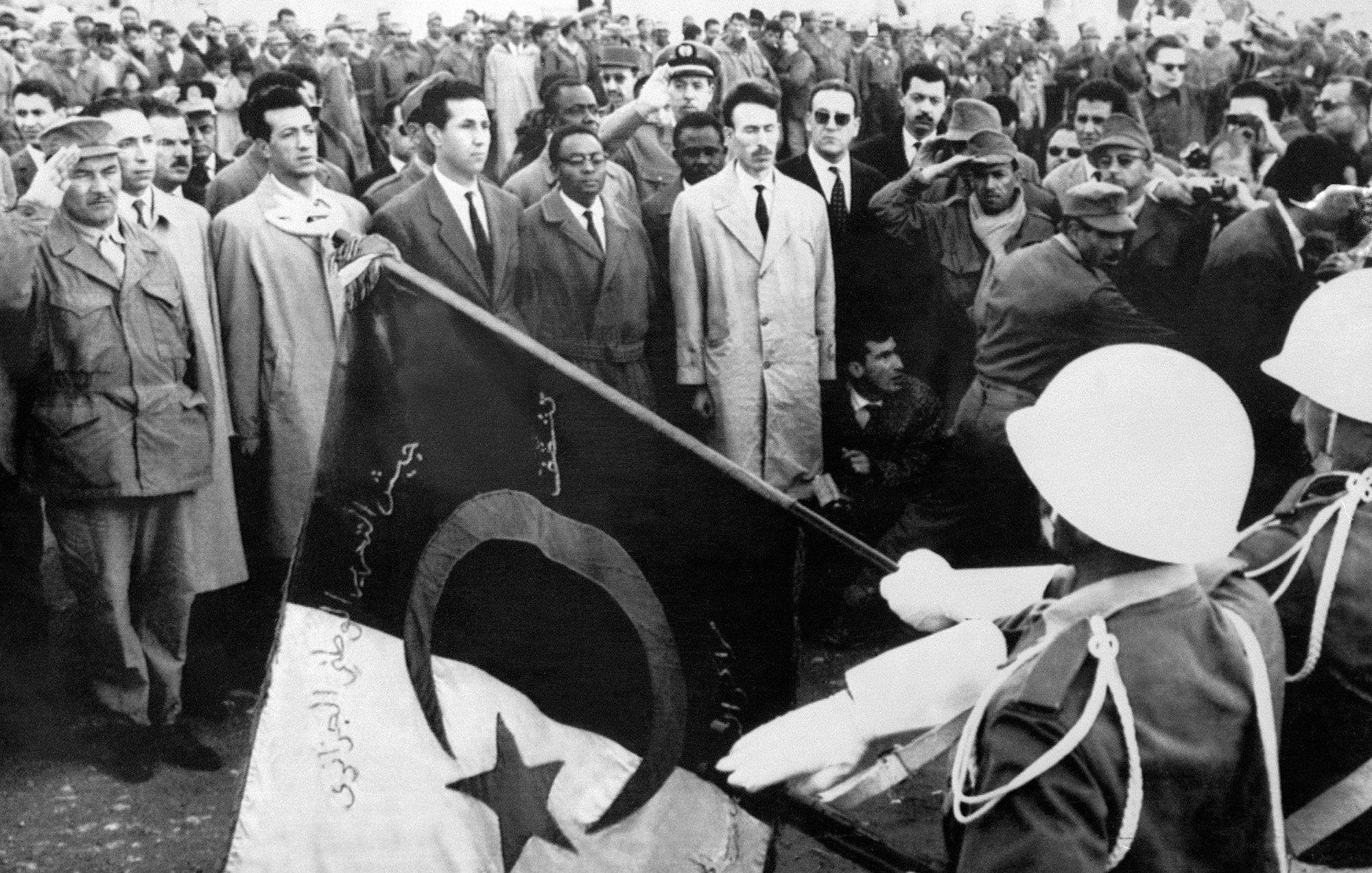 The image depicts a historical scene with a large crowd gathered, likely for a ceremonial or significant event. In the foreground, two soldiers are saluting while holding a flag, which features a crescent and a star, elements often associated with some national flags. The crowd appears to consist of men in various types of clothing, with some wearing military uniforms and others in suits. The overall atmosphere suggests a moment of national importance, possibly related to independence or a political event.
