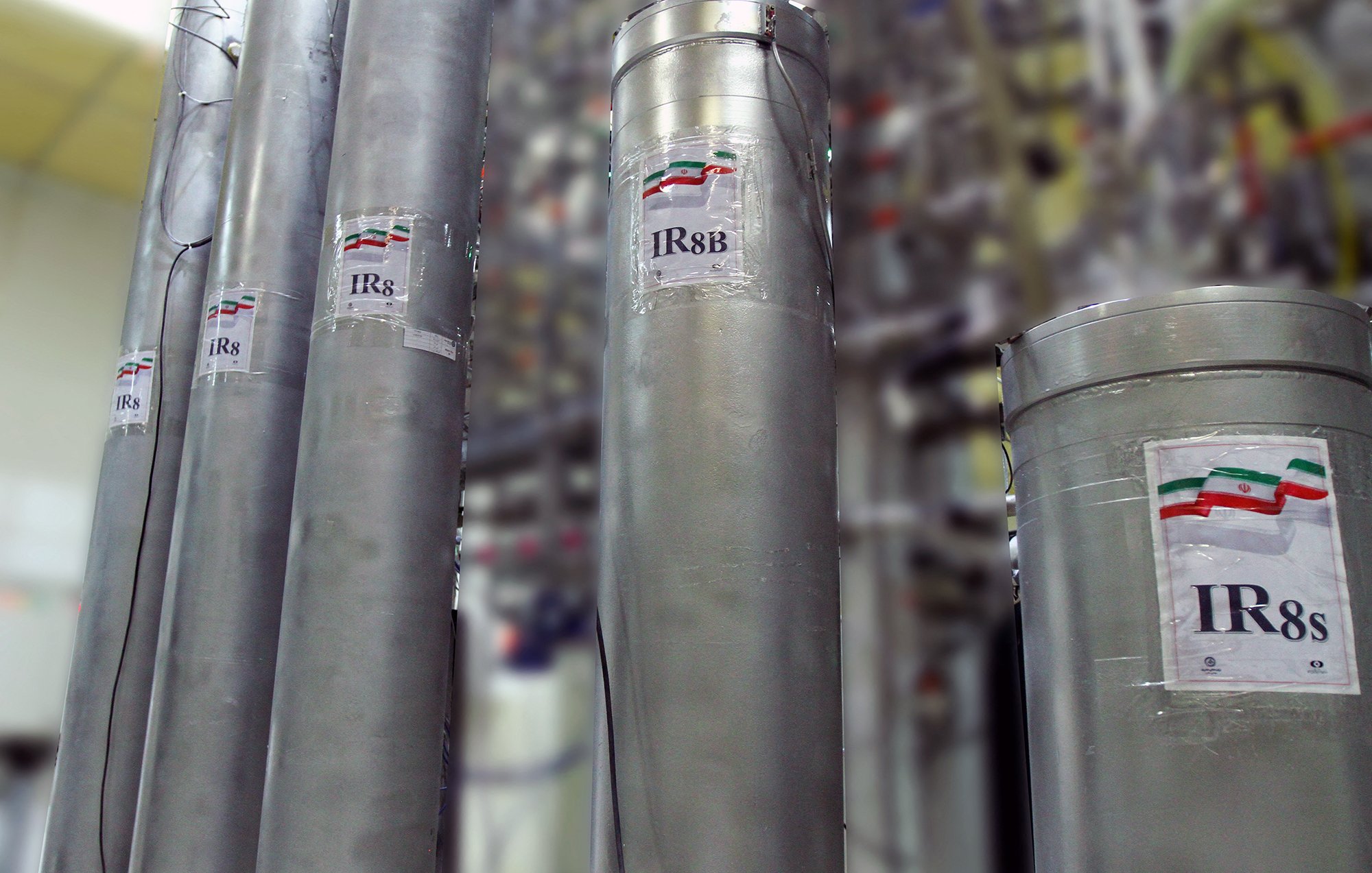 The image features tall, cylindrical metal containers labeled with the designation "IR8" and "IR8s," which suggests they are related to technological or industrial equipment, possibly in a laboratory or manufacturing setting. There are also labels indicating a flag, likely representing a country, next to the designations. The background is blurred, showing a complex arrangement of machinery or similar structures, indicating a sophisticated environment, possibly related to nuclear technology or similar fields.