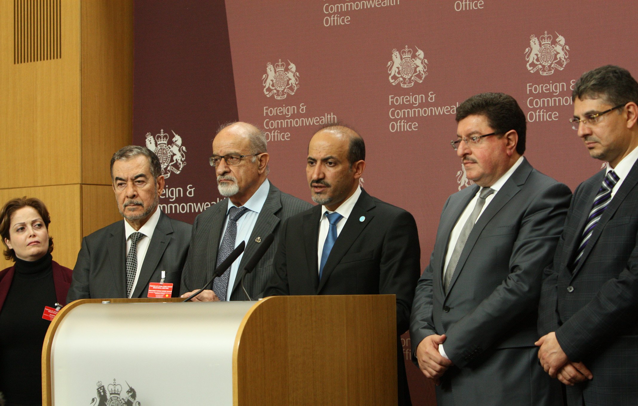 L'image montre un groupe de plusieurs personnes lors d'une conférence de presse. Elles se tiennent devant un podium avec un emblème officiel, et une toile de fond affichant "Foreign & Commonwealth Office". Les individus paraissent sérieux et concentrés, probablement en train de faire une déclaration ou de répondre à des questions. Les personnes sont habillées de manière formelle, illustrant l'importance de l'événement.