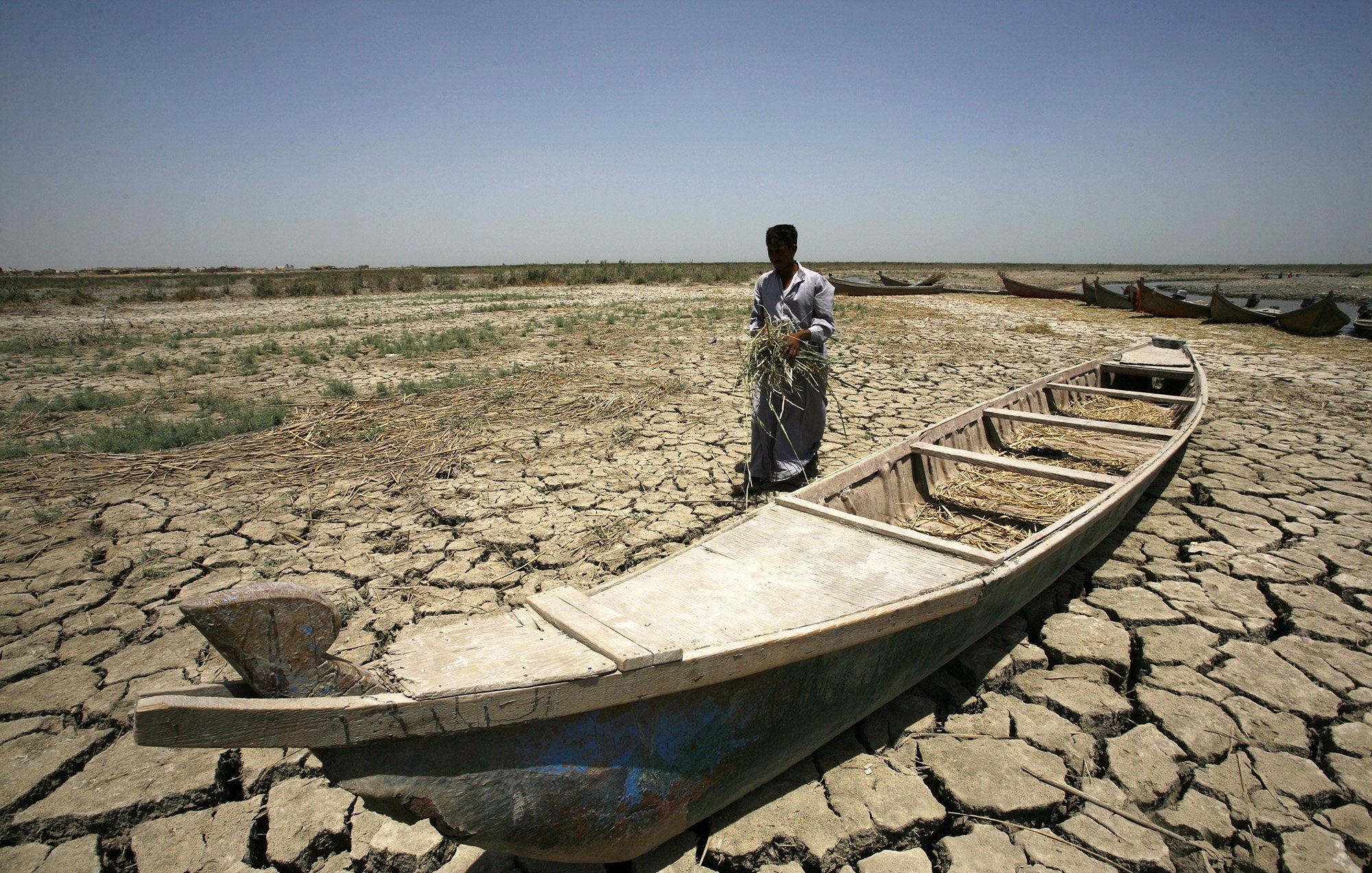 L'image montre un homme debout sur un sol craquelé, typique d'une région aride. Il semble tenir une poignée de végétation, peut-être de l'herbe ou des tiges. À ses pieds se trouve une barque vide, qui est en partie enfouie dans le sol sec. En arrière-plan, on peut apercevoir d'autres barques, mais le paysage aride, sans présence significative d'eau, souligne un environnement désolé et dégradé. Le ciel est dégagé et lumineux, renforçant l'atmosphère de chaleur et de sécheresse.