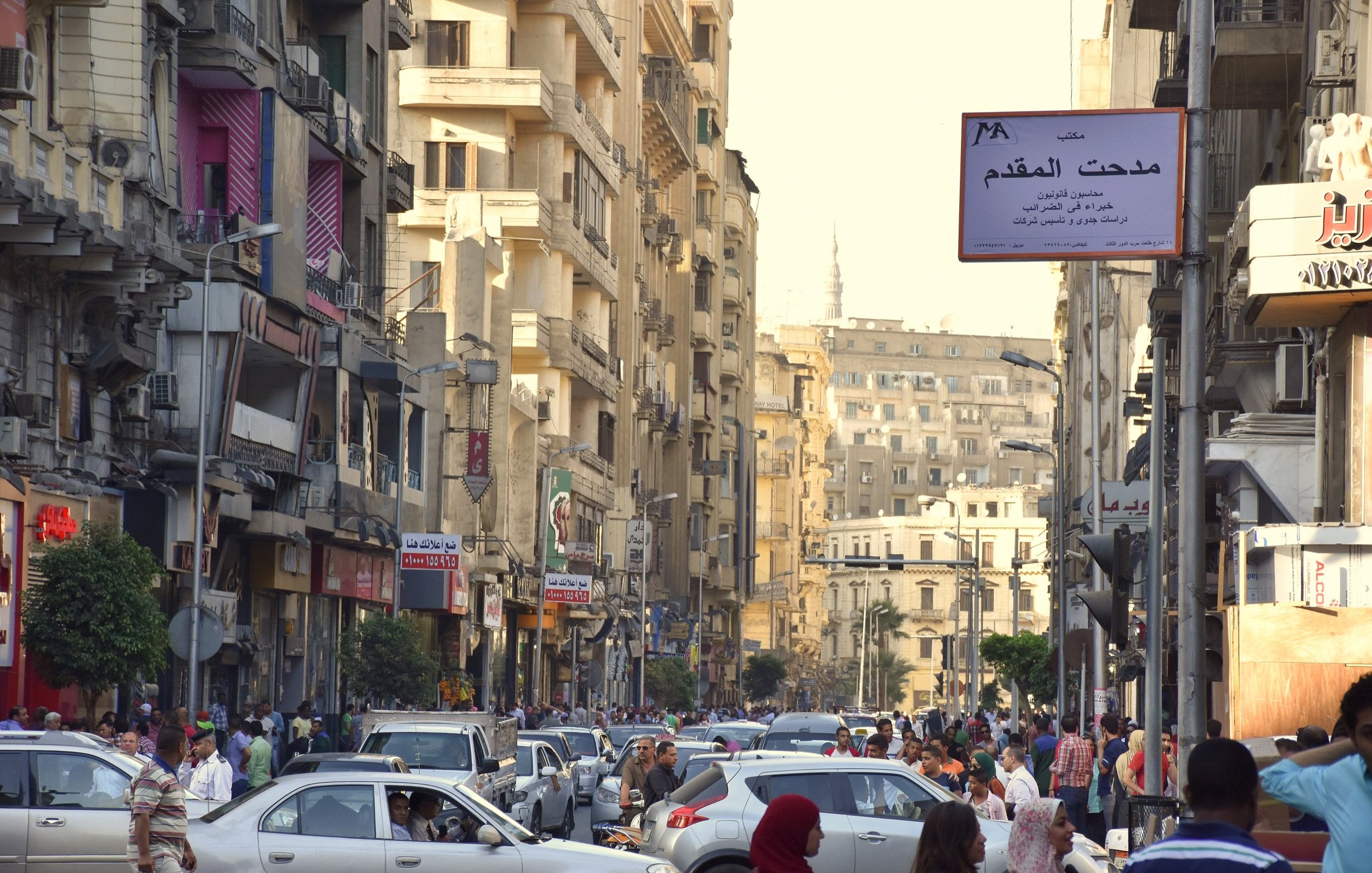 L'image montre une rue animée remplie de voitures et de passants. On peut voir des bâtiments de plusieurs étages de part et d'autre de la rue, avec des balcons et des façades variées. La scène semble se dérouler pendant la journée, la lumière du soleil illuminant l'environnement urbain. Des panneaux de signalisation et des enseignes en arabe sont visibles, ajoutant à l'atmosphère locale de cette ville. Les gens semblent se déplacer rapidement, créant une ambiance dynamique et vivante.