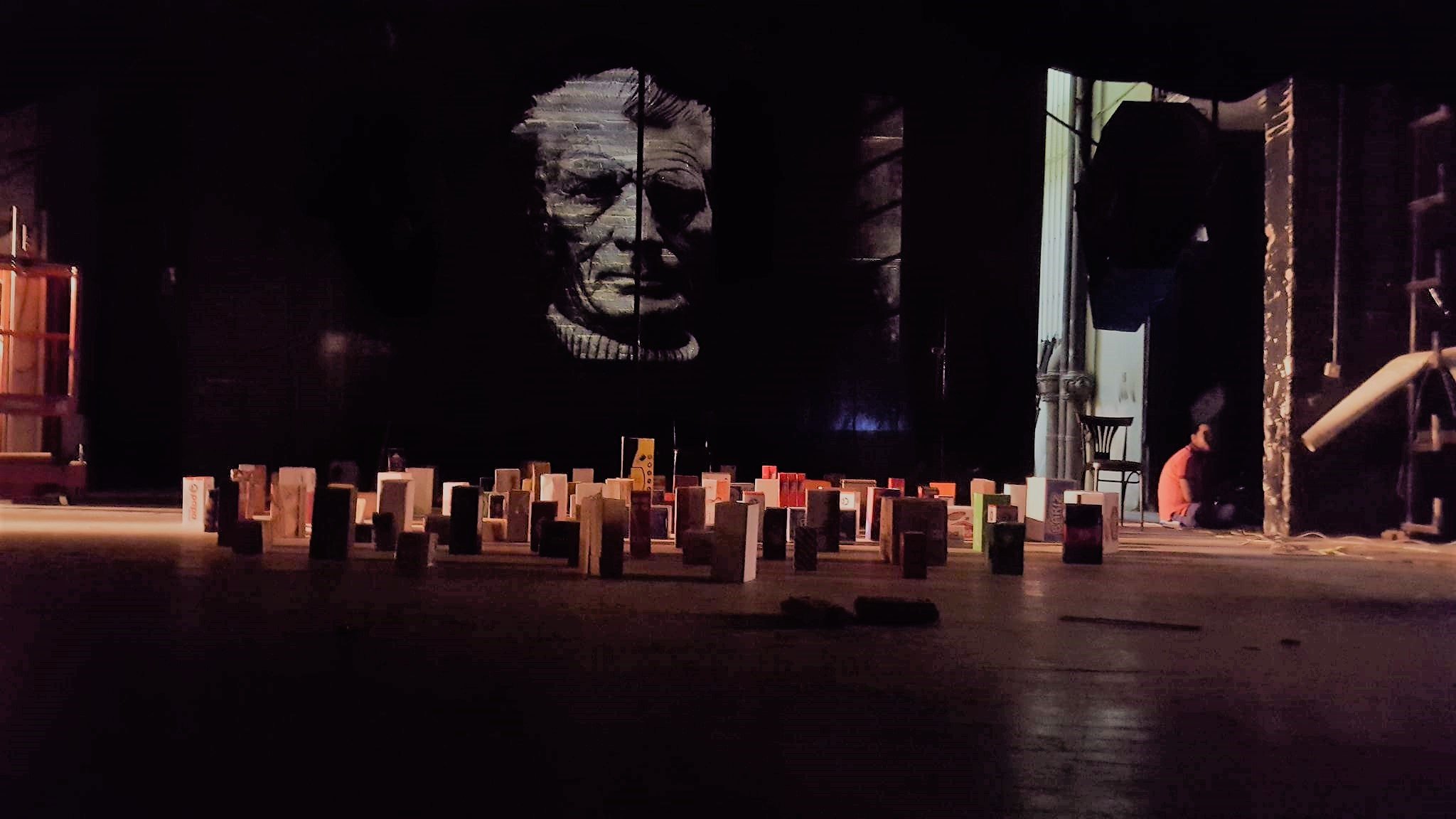 L'image montre une scène intérieure avec un grand mur sombre sur lequel se trouve un portrait en noir et blanc d'une personne âgée. Au premier plan, il y a un sol rempli de petites structures ressemblant à des livres ou des modules disposés de manière organisée. À l'arrière-plan, on peut apercevoir une personne assise, probablement en train de travailler ou de se préparer. L'éclairage est faible, créant une atmosphère mystérieuse.