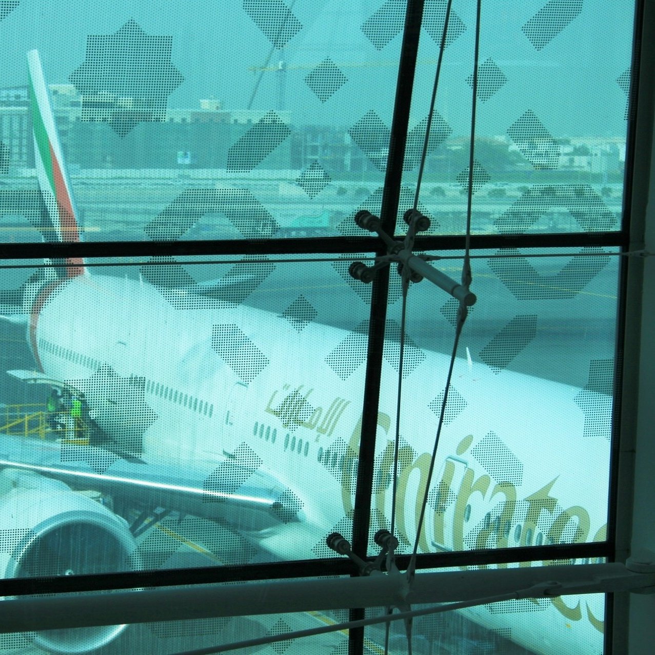 در این تصویر، یک هواپیمای متعلق به شرکت هواپیمایی امارات در حال ایستادن در فرودگاه دیده می‌شود. نمای تصویر از داخل یک ساختمان فرودگاهی است و بخشی از شیشه‌های پنجره با الگوهای خاصی پوشانده شده است. هواپیما در حال آماده‌سازی برای پرواز است و تعدادی از کارکنان روی نردبان ایستاده‌اند تا کارها را انجام دهند. در پس‌زمینه، ساختمان‌ها و تجهیزات فرودگاهی نیز قابل مشاهده هستند. جو عمومی تصویر کمی مه‌آلود به نظر می‌رسد.