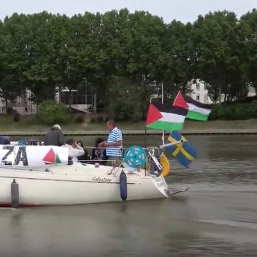 The image shows a boat on a body of water with several individuals on board. The boat has a prominent banner that reads "SHIP TO GAZA," and it is adorned with various flags, including the Palestinian flag and the flag of Sweden. In the background, there are trees and buildings along the shoreline. The scene conveys themes of solidarity and activism related to Gaza.