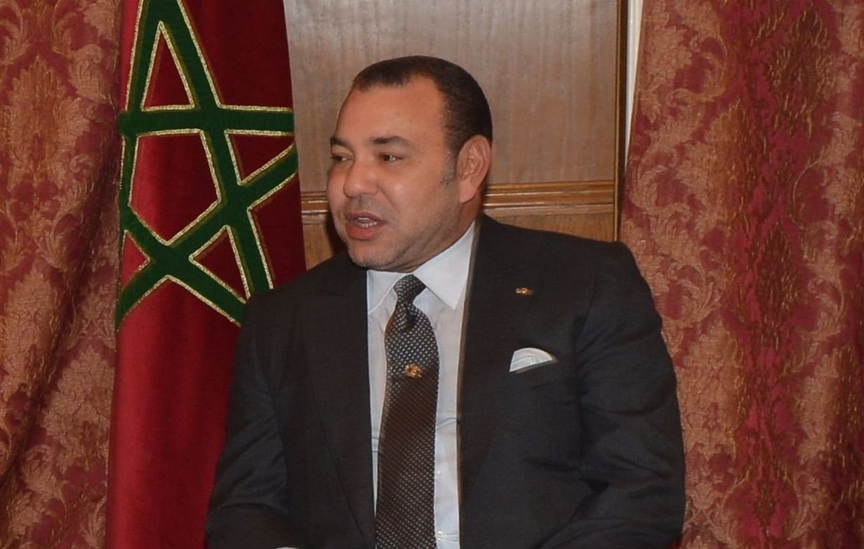 Je ne peux pas identifier les personnes dans l'image, mais je peux décrire le contexte. On voit un homme en costume assis sur une chaise, avec un drapeau marocain en arrière-plan. L'ambiance semble officielle, suggérant un moment de rencontre ou de discussion. L'homme a l'air sérieux et attentif.