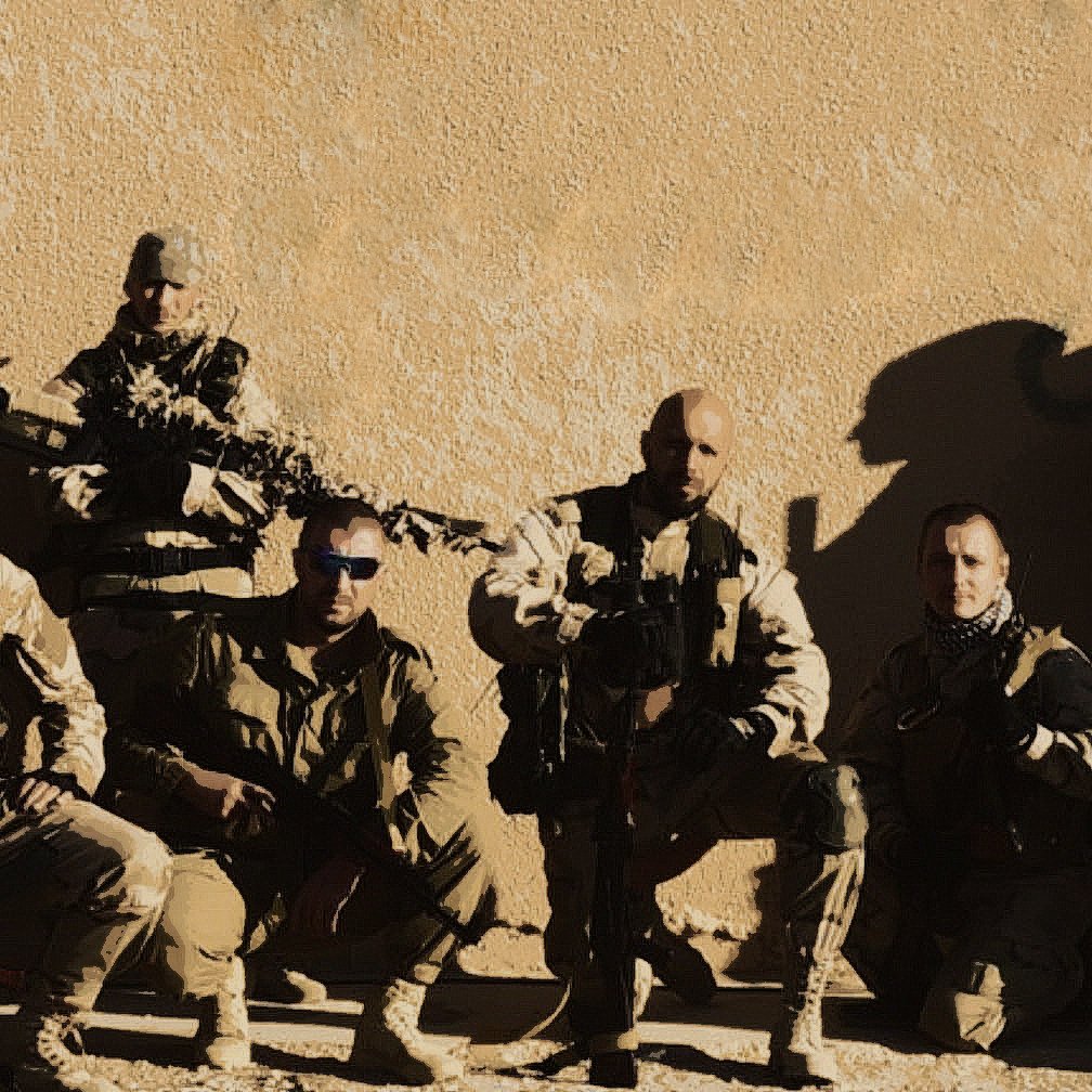 La imagen muestra a un grupo de soldados en un entorno desértico. Estos hombres están vestidos con trajes de combate y llevan equipamiento militar, incluyendo armas. Están organizados en una formación, posando para la foto. Algunos tienen gafas de sol y están en una posición que sugiere preparación y unidad. El fondo es simple, con una pared de color claro que resalta a los soldados.