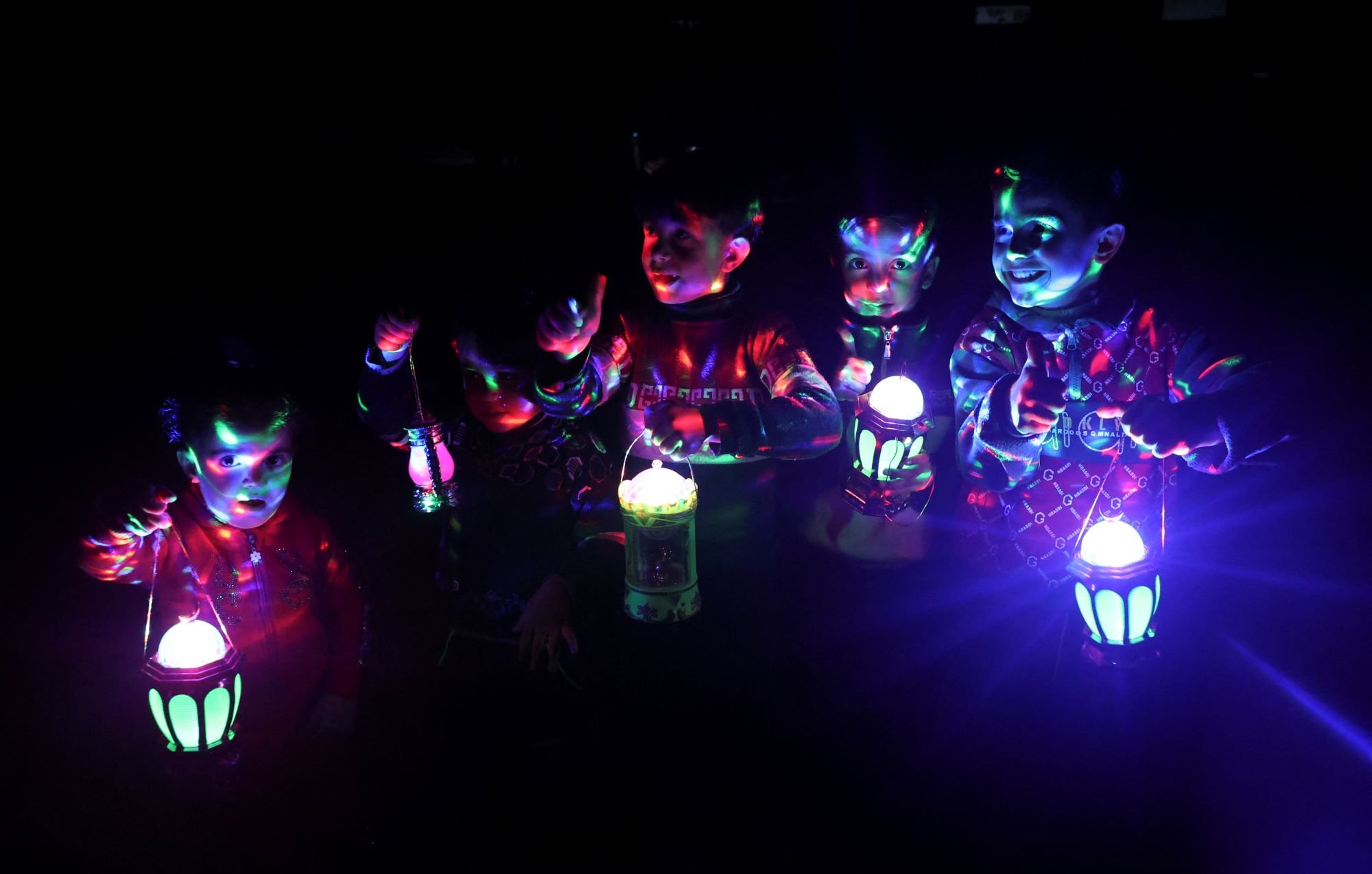 L'image montre un groupe d'enfants dans l'obscurité, tenant des lanternes colorées qui émettent des lumières vives. Les lumières créent des reflets et des motifs multicolores sur leurs visages et leurs vêtements. Les enfants semblent joyeux et amusés, s'amusant avec les lanternes pendant la nuit. L'ambiance est festive et magique grâce aux jeux de lumière.