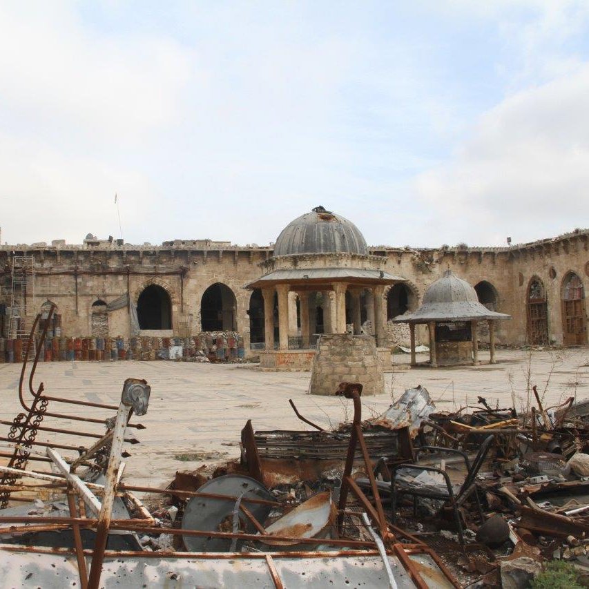 L'image montre un site historique en ruines. On peut voir une cour spacieuse entourée de murs en pierre, avec des arches et des colonnes qui témoignent d'une architecture ancienne. Au centre, il y a deux structures en forme de dômes. Le sol est recouvert de débris métalliques et de morceaux de ferraille, suggérant que le lieu a été touché par des événements destructeurs. Le ciel est nuageux, ajoutant à l'atmosphère de désolation.