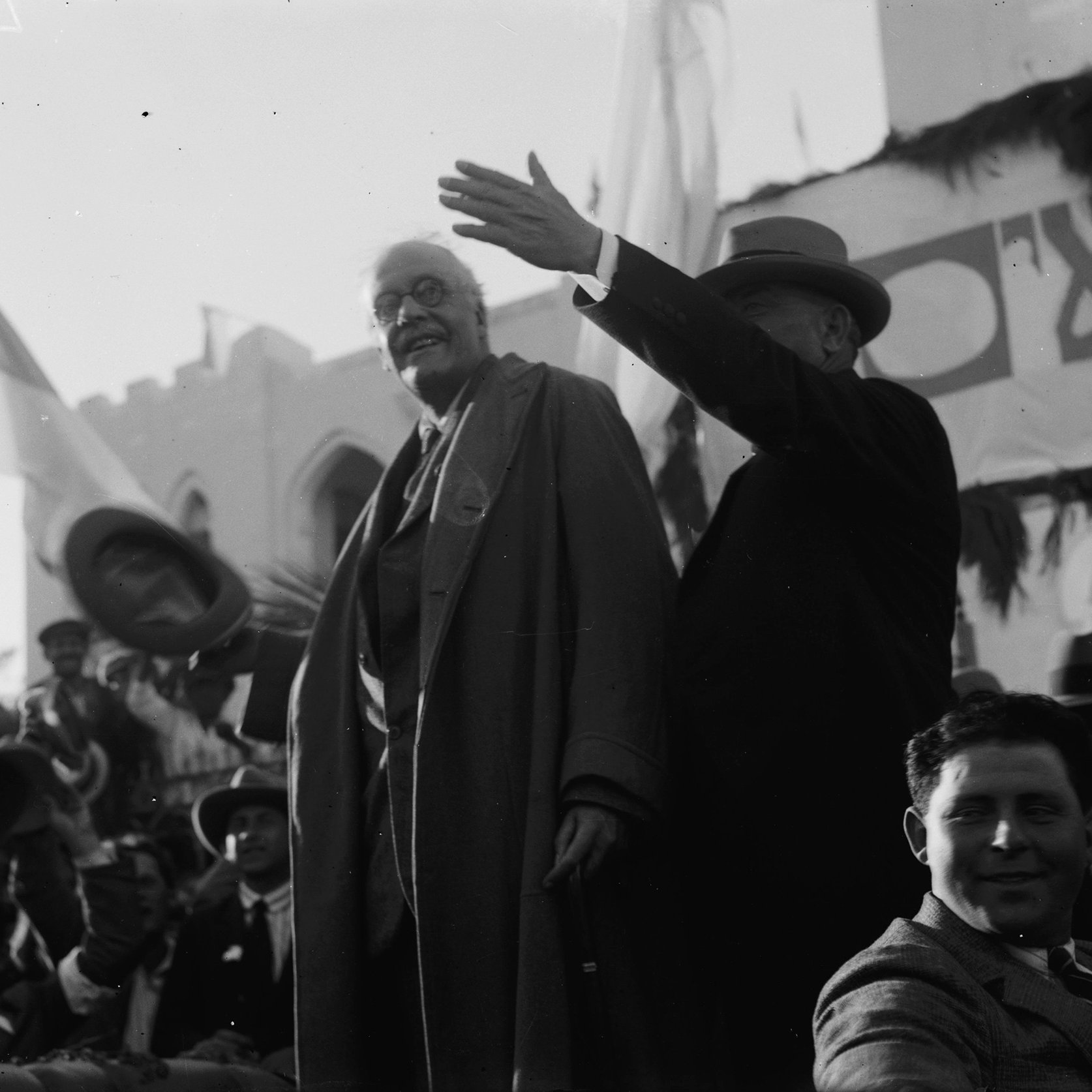 در این تصویر، دو مرد در حال سخنرانی یا معرفی هم دیگر هستند. یکی از آن‌ها به وضوح در حال صحبت کردن است و دستش را به علامت اشاره به سمت جمعیت بالا برده است. مرد دیگر کنار او ایستاده و به نظر می‌رسد که به جمعیت توجه دارد. پس‌زمینه تصویر ازدحام جمعیت را نشان می‌دهد که در حال تماشای آنها هستند. بعضی پرچم‌ها و طراحی‌هایی در پس‌زمینه دیده می‌شوند که احتمالاً به یک رویداد خاص اشاره دارند. این تصویر حال و هوای تاریخی و اجتماعی را به نمایش می‌گذارد.