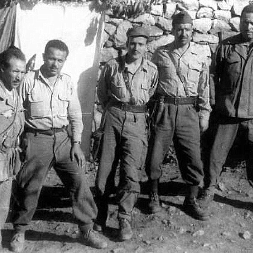 The image shows a group of five soldiers standing together in a rural setting. They are wearing military uniforms and appear to be posing for the photo. The background consists of a stone wall and a makeshift tarp or cloth, suggesting a temporary encampment. The soldiers have serious expressions, reflecting the context of their environment. Overall, the image conveys a sense of camaraderie among them in a military setting.