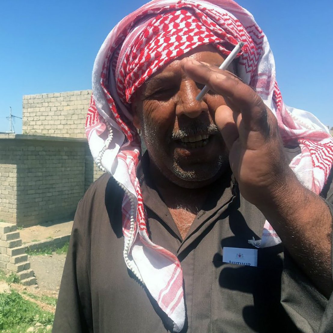 The image shows a man wearing a traditional headscarf, often referred to as a keffiyeh or ghutrah, with a red and white pattern. He is standing outdoors, possibly in a rural area, with stone structures visible in the background. The man appears to be smiling or in a candid moment, holding a cigarette between his fingers. The setting is bright, indicating a sunny day, and the landscape around him includes some greenery and dirt paths.
