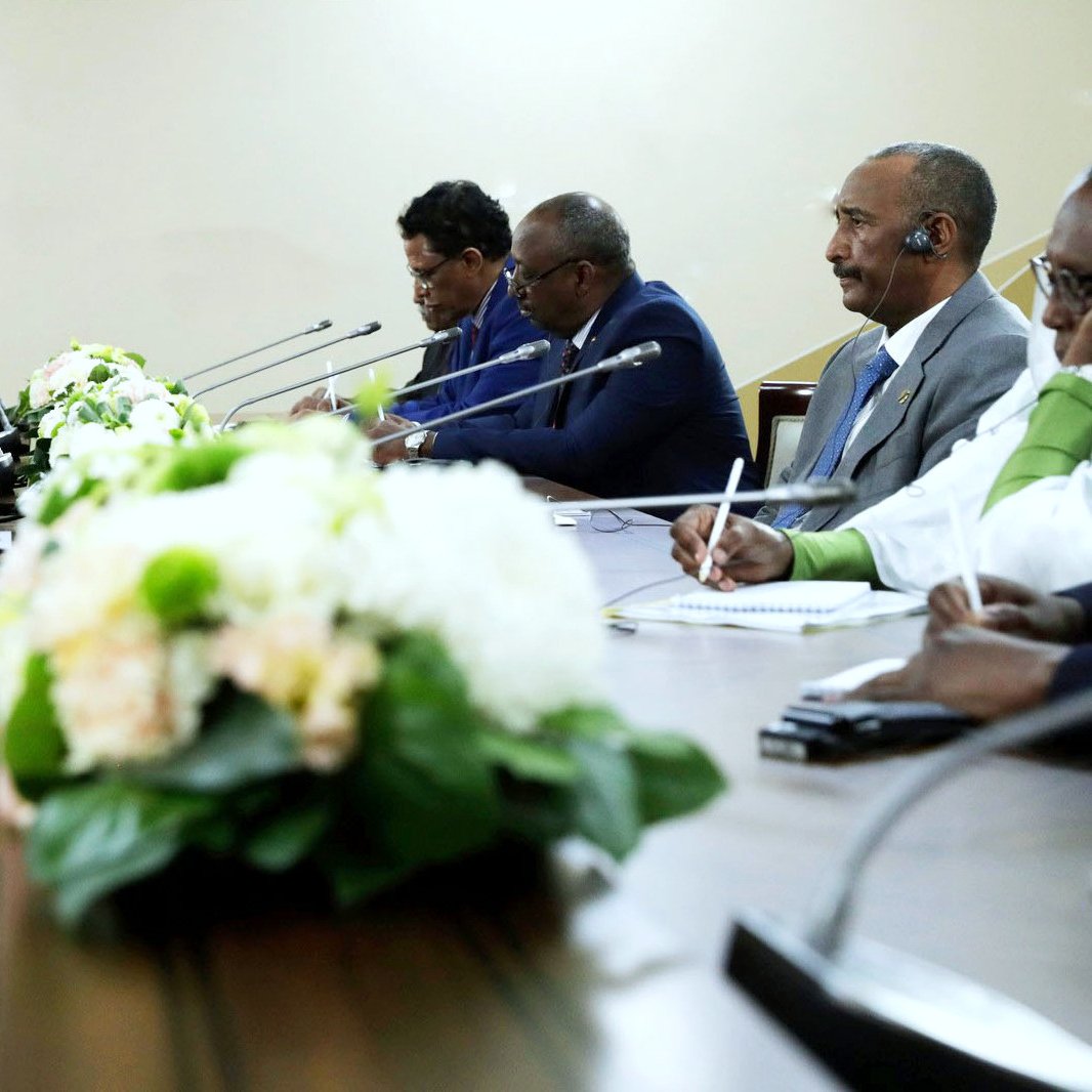 در تصویر یک جلسه رسمی دیده می‌شود که چندین نفر در دو طرف میز نشسته‌اند. در وسط میز، گل‌های زیبایی قرار دارند که فضا را تزئین کرده‌اند. افراد در حال گفتگو هستند و تجهیزات صوتی نیز روی میز قرار دارد. به نظر می‌رسد موضوع جلسه مهم و رسمی است و افراد با تمرکز در حال تبادل نظر و بحث هستند.