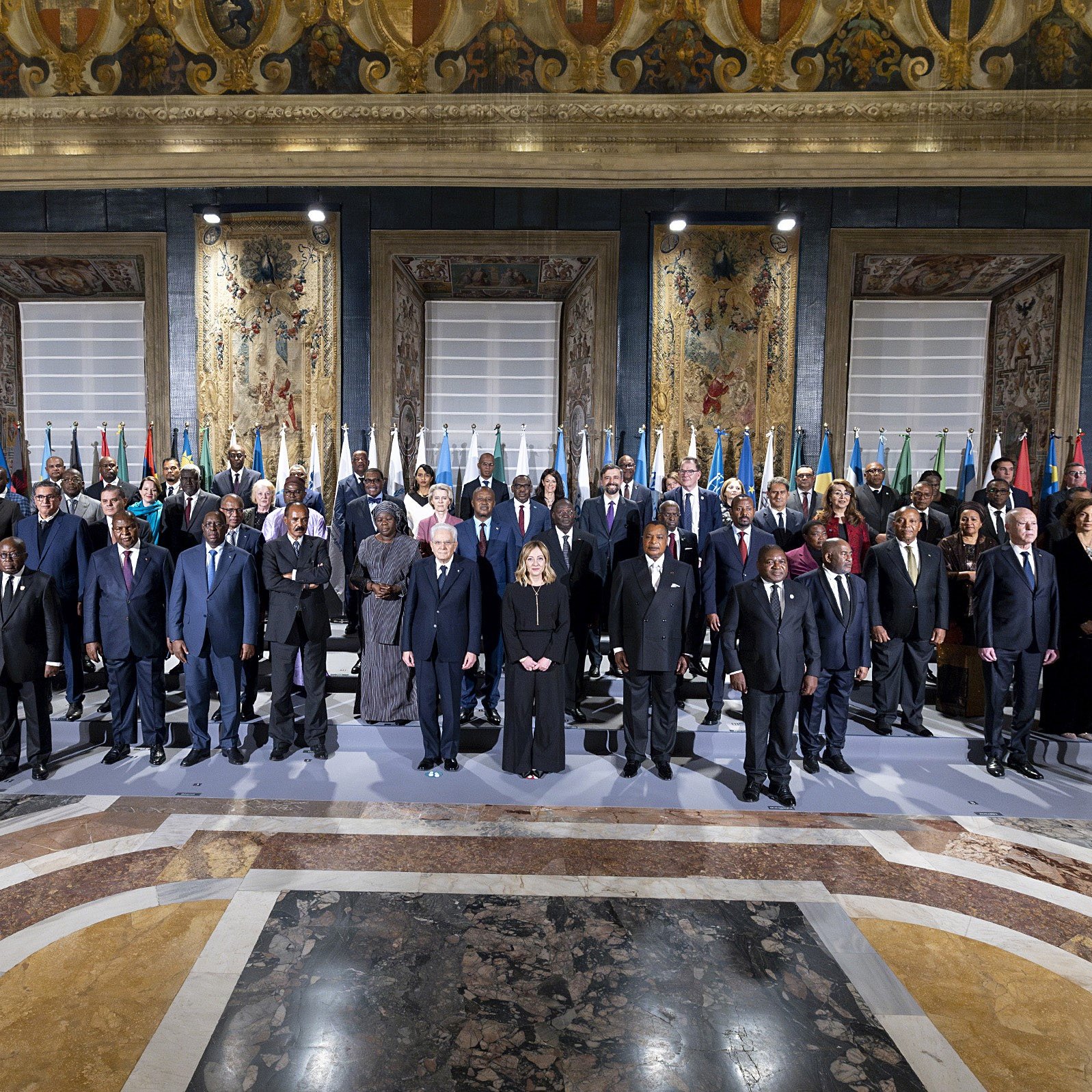 L'immagine mostra un gruppo di persone in un ambiente ufficiale, probabilmente durante un incontro o una conferenza internazionale. Si vedono diversi uomini e donne in abiti formali, che rappresentano vari paesi, dato che ci sono bandiere nazionali esposte sullo sfondo. L'atmosfera sembra seria e solenne, con tutti i partecipanti schierati in modo ordinato. Le decorazioni e l'architettura dell'ambiente circostante danno un tocco di grandezza all'evento.