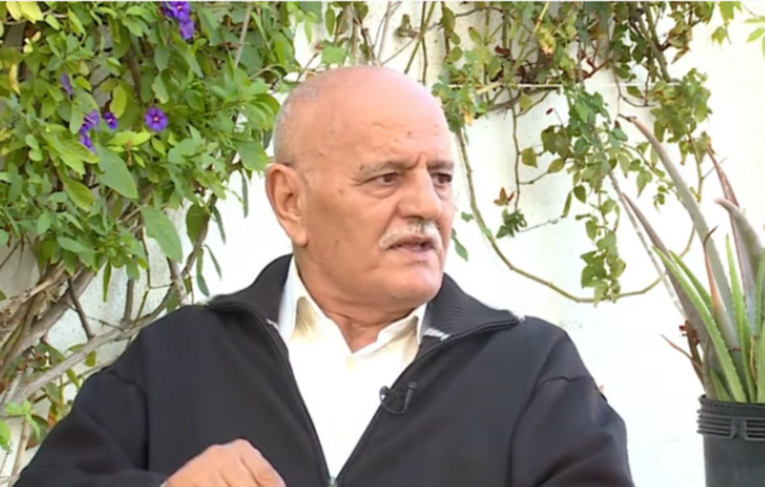 تظهر الصورة شخصًا في منتصف العمر يجلس في مكان خارجي، محاطًا بالنباتات الخضراء. يرتدي سترة داكنة ولديه لحية وشعر رمادي. يبدو وكأنه يتحدث أو يعبّر عن رأيه في موضوع ما، حيث يرفع إصبعه للتأكيد على نقطة سابقة. الخلفية تحتوي على نباتات وأزهار، مما يضفي جوًا طبيعيًا على المشهد.