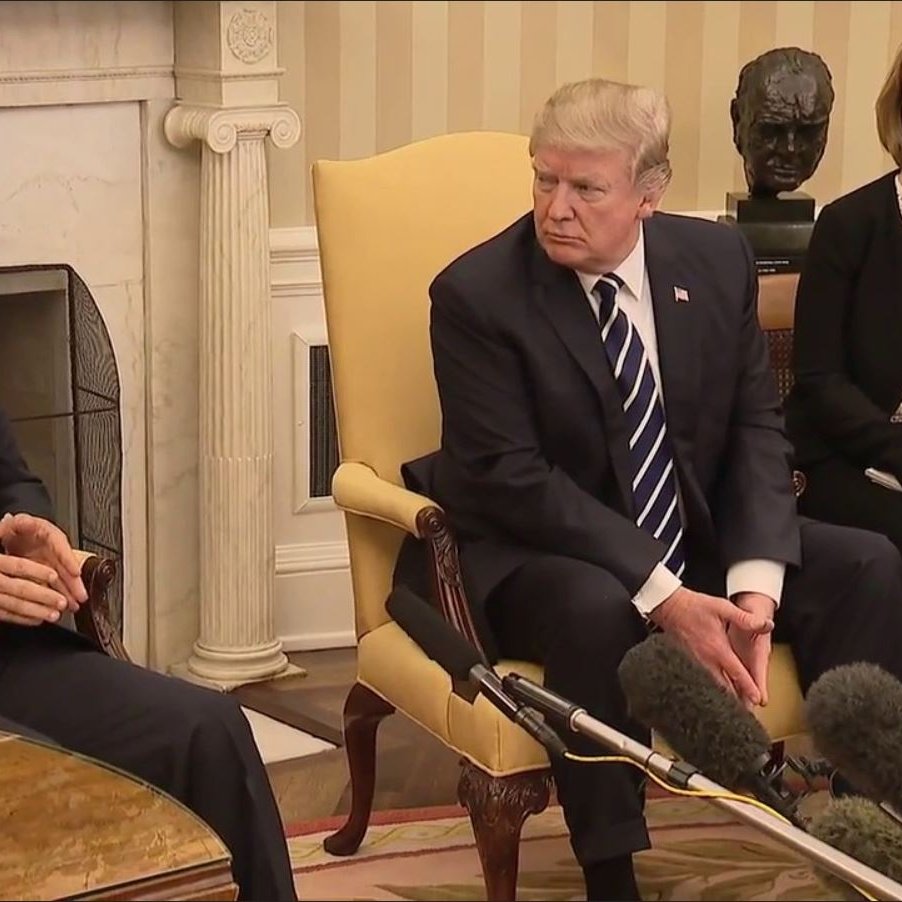 The image shows two men sitting in a formal setting, likely a meeting room in a government building. One man, on the left, is wearing a dark suit and a red tie, appearing to speak or make a point. The other man, on the right, is dressed in a dark suit with a striped tie and is attentively listening or observing. There are microphones positioned between them, indicating that their conversation is being recorded or broadcast. The background features a light-colored wall and a decorative element, possibly a bust or sculpture, along with a warm lamp on the table.