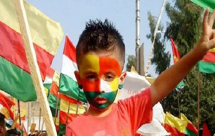 L'image montre un jeune garçon au milieu d'une manifestation ou d'une célébration. Il a le visage peint aux couleurs du drapeau, probablement inspirées par les couleurs du Cameroun, avec des nuances de vert, rouge et jaune. Il porte un t-shirt rouge et affiche un signe de paix avec sa main, tout en étant entouré de drapeaux aux couleurs similaires. L'atmosphère semble festive et engagée.