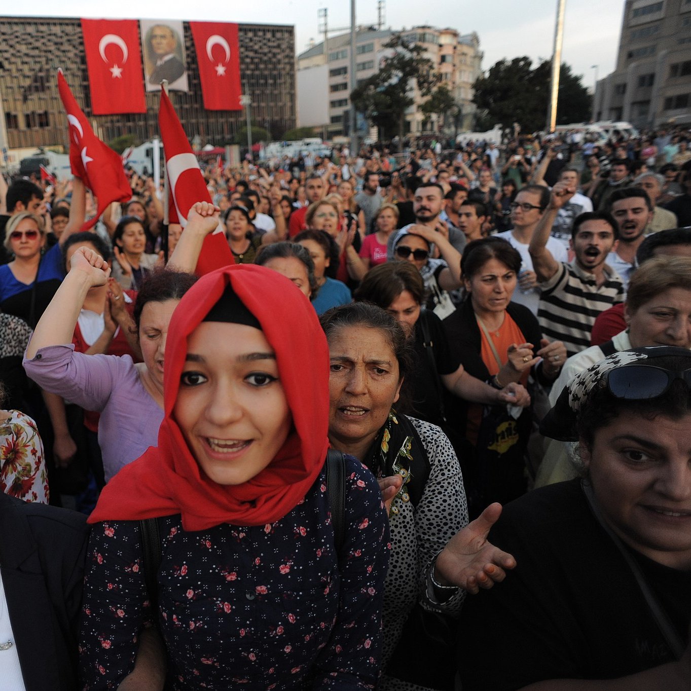 تظهر في الصورة حشود من الناس تجمعوا في مكان واحد، حيث يحمل بعضهم الأعلام التركية. يبدو أن هناك مشاعر حماسية واضحة بين المتظاهرين، حيث يرفعون أصواتهم ويعبرون عن آرائهم. ترتدي بعض النساء الحجاب، بينما تظهر في المقدمة فتاة صغيرة ترتدي غطاء رأس. الخلفية تضم مباني وبعض الصور التي تعكس أجواء الاحتجاج أو الاحتفاء.